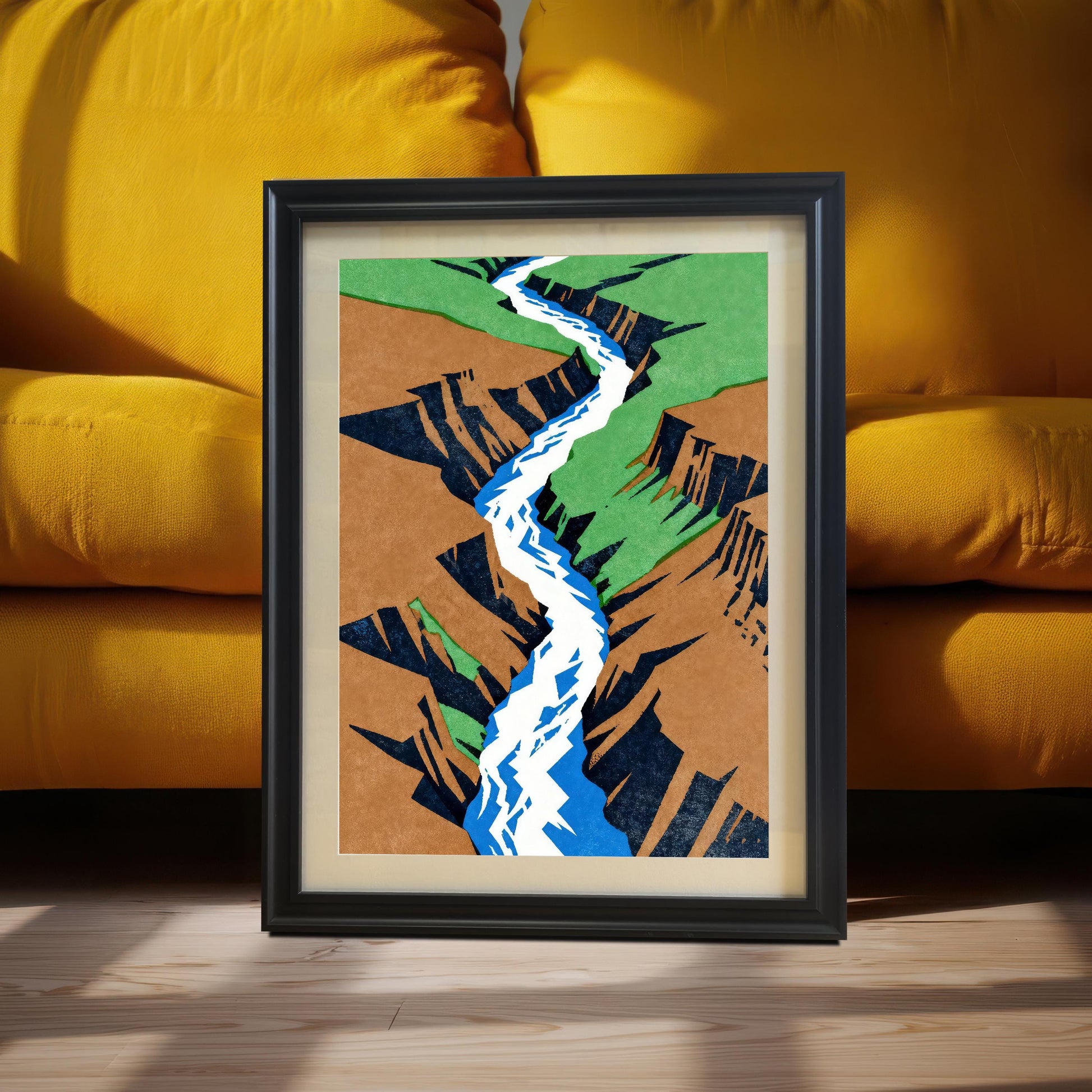 A black-framed landscape art print with a cream mount, leaning against a vibrant mustard yellow sofa on a light wood floor, captured in warm, natural sunlight.