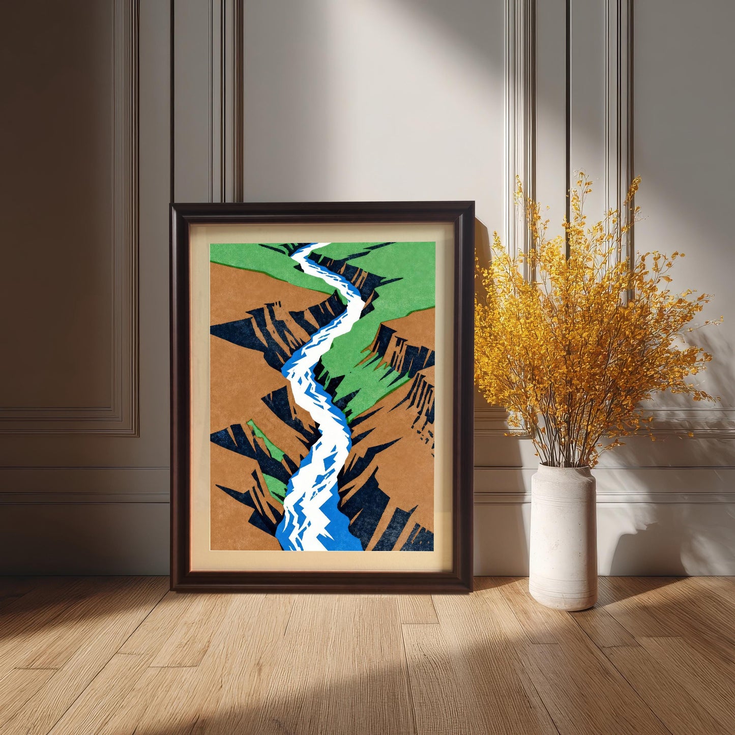 A dark coffee-colored wood frame with a cream mount, showcasing the colorful canyon art as it sits on the floor next to a tall vase of bright yellow wildflowers in a sophisticated, sun-drenched room.