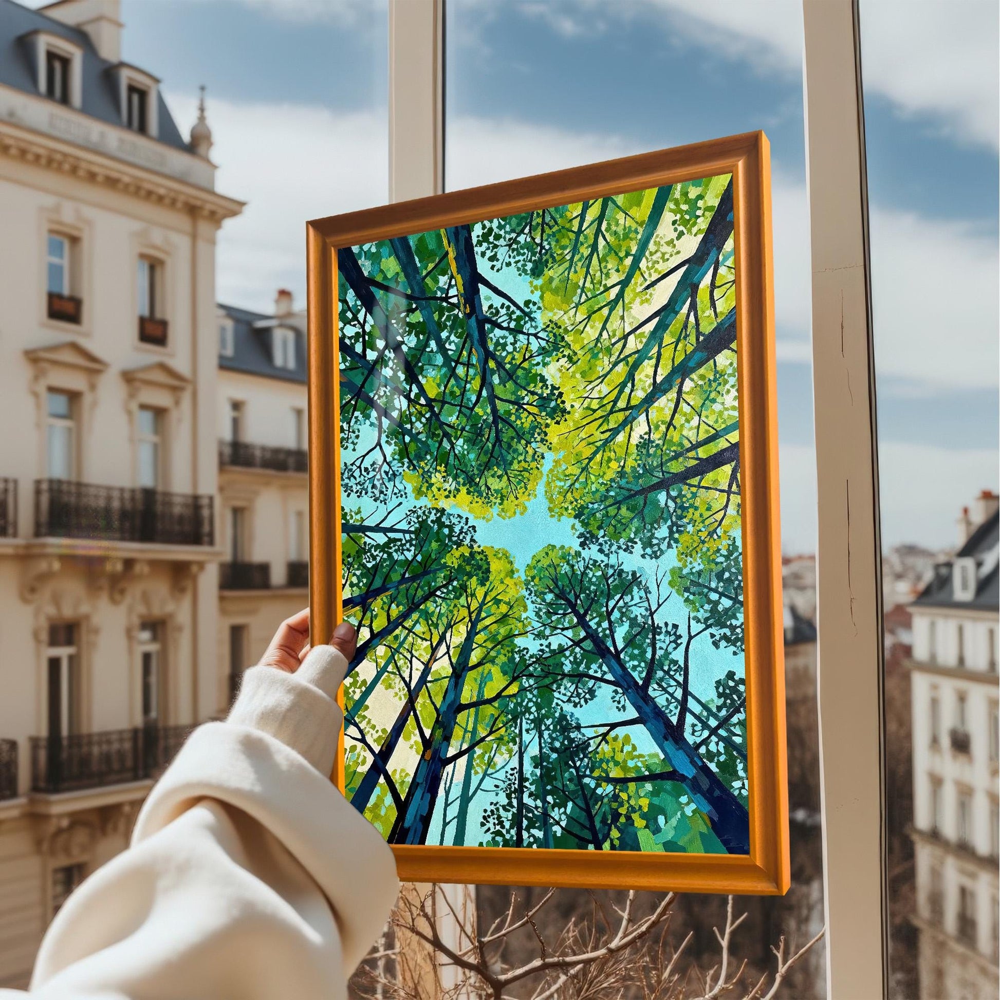 A hand holding a framed forest canopy art print up against a window with a blurred European city street and blue sky in the background.