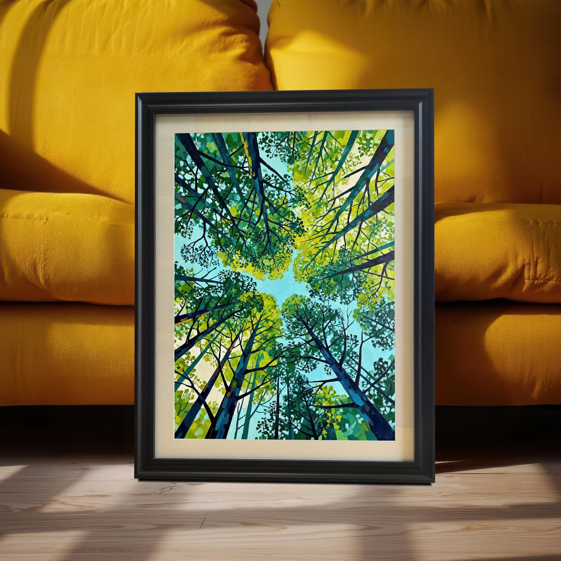 Close-up of a matted nature art print in a black wood frame, resting on a wooden floor against the cushions of a vibrant yellow sofa.