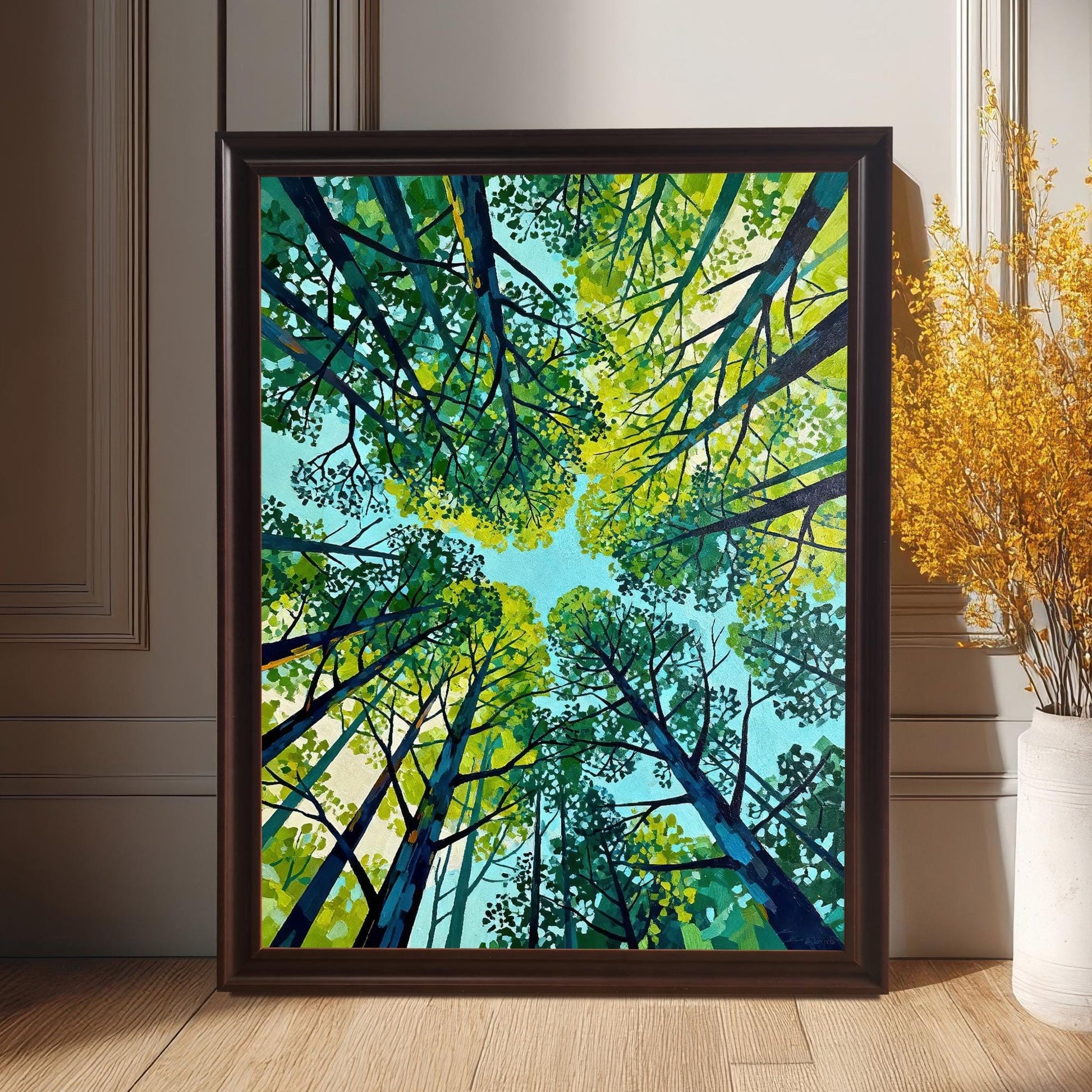 Vertical wall art of a green forest canopy in a dark coffee-colored wood frame leaning against a wall next to a vase of yellow flowers.