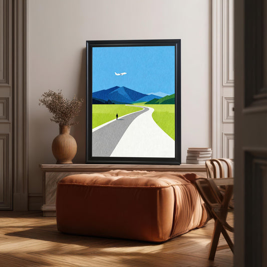 A large black framed minimalist landscape print featuring a blue sky, mountains, and a winding road, styled leaning on a wooden sideboard in a sunlit, modern interior.