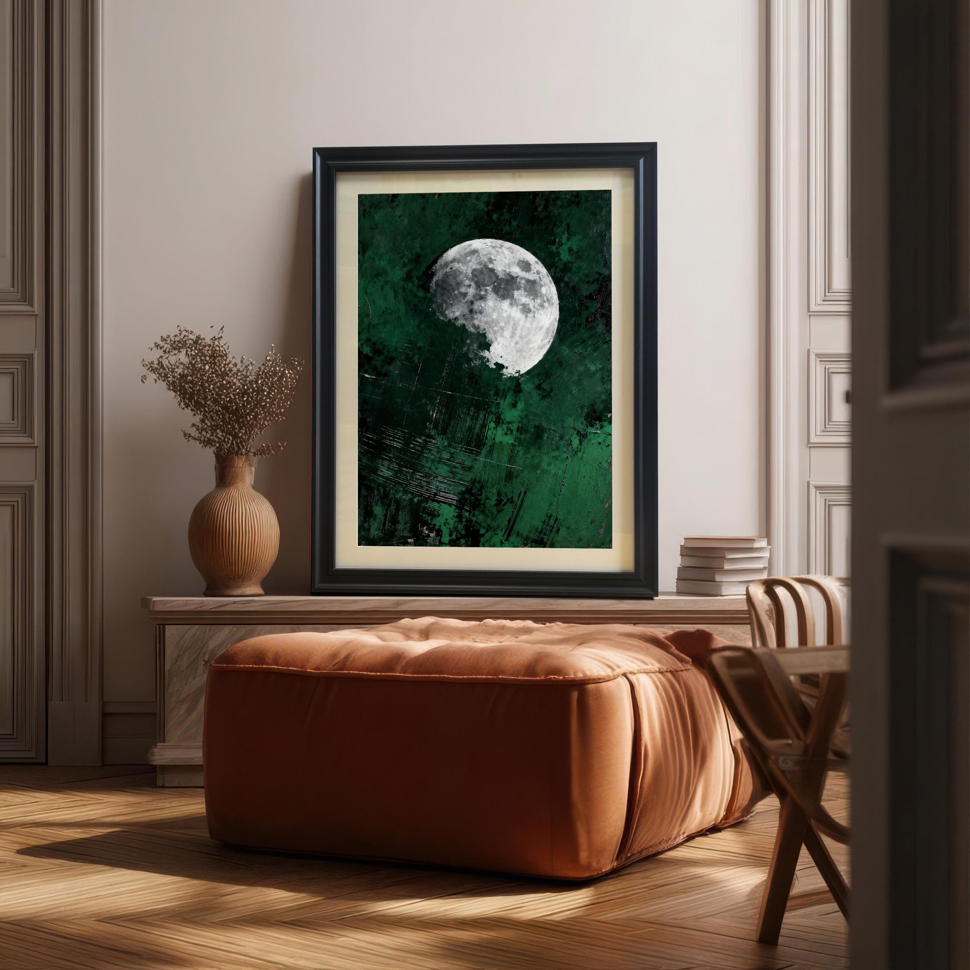 Large black framed moon wall art with an off-white mat board leaning on a wooden console table in a modern, sunlit living room featuring a tan leather ottoman.