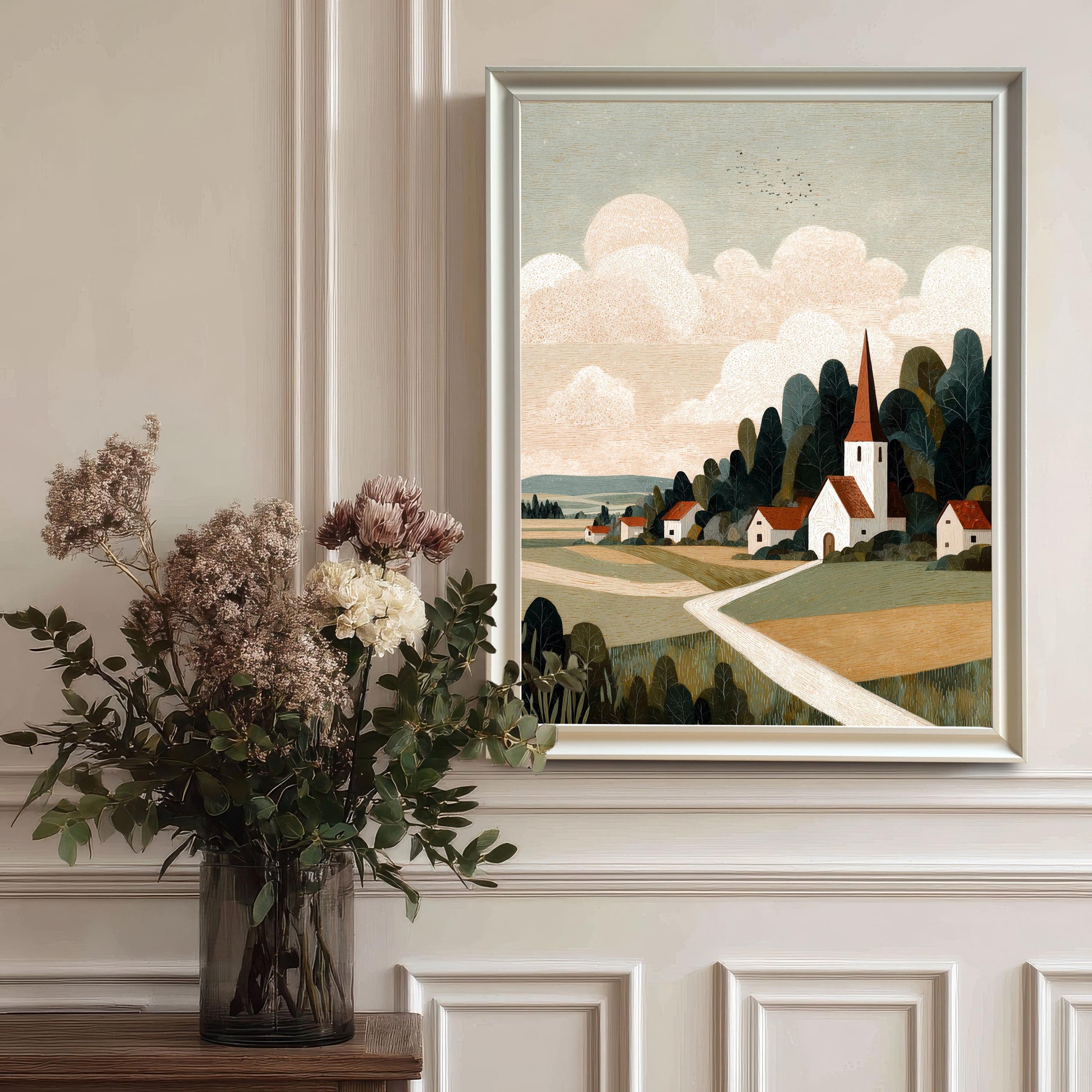 A white-framed landscape art print of a rural village with a church spire, hanging on a white wall with classic molding and styled with a large floral vase in the foreground.