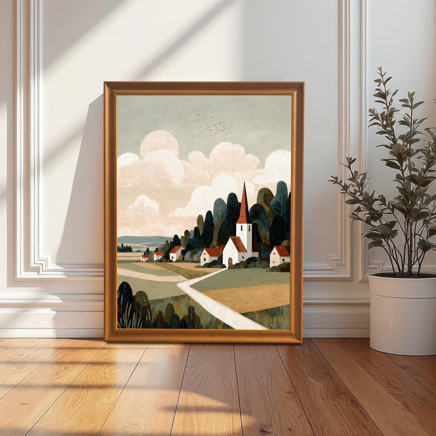 A wood-framed landscape art print of a village and church leaning against a white wall with elegant molding on a hardwood floor.