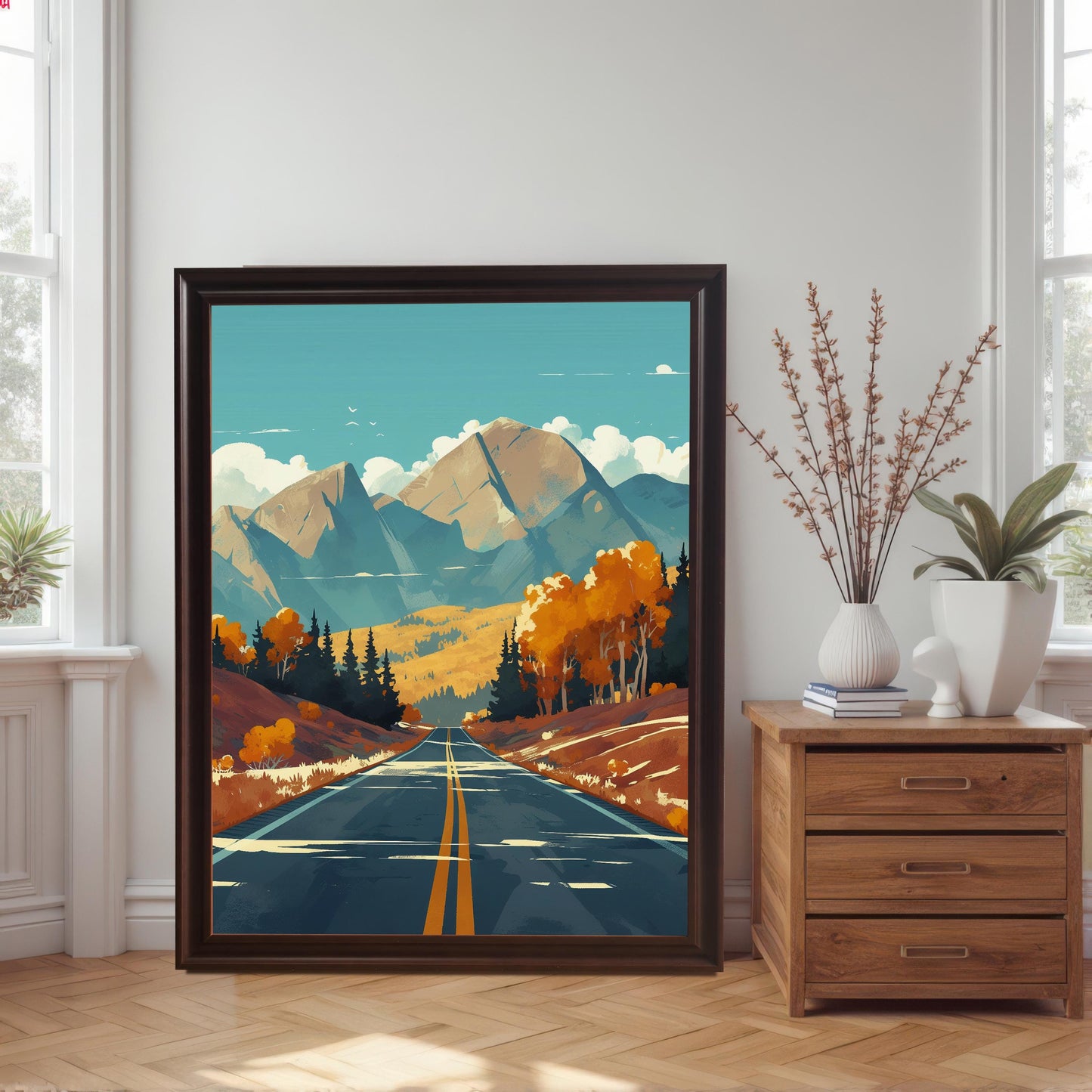 Large-scale coffee wood framed art print of a scenic mountain highway, leaning against a white wall next to a wooden dresser in a bright room with herringbone floors.