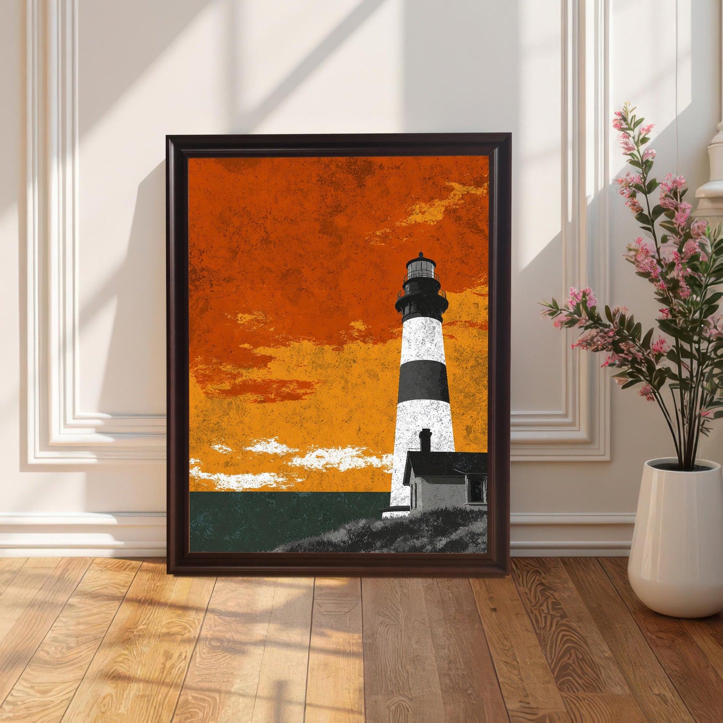 A coffee brown wood-framed lighthouse art print sitting on a polished hardwood floor against a white paneled wall, styled with a white vase of pink flowers.