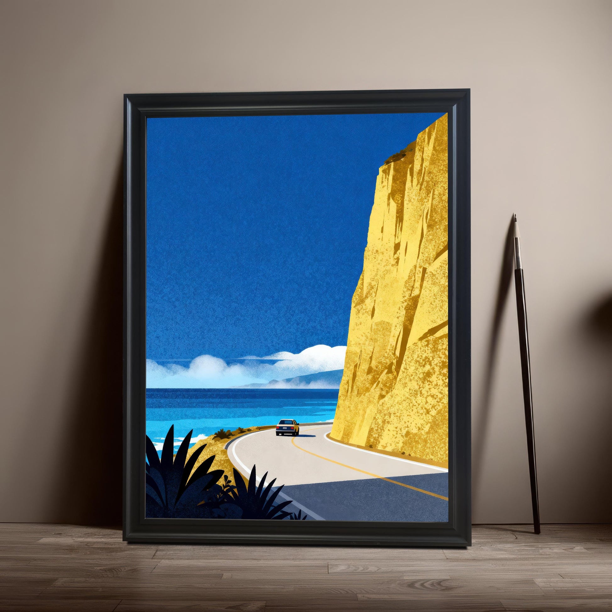 A vertical black wood-framed art print of a coastal highway scene leaning against a neutral wall on a wooden floor, next to a long artist's brush.
