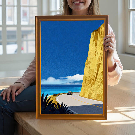 A woman holding an orange-toned wood-framed coastal art print in front of her while sitting at a light wood table in a sunlit room.
