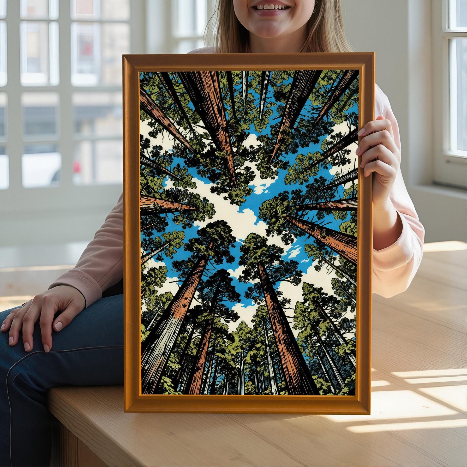A woman holding a large orange wood framed art print of a sequoia forest to show scale, sitting in a bright, sun-drenched room.