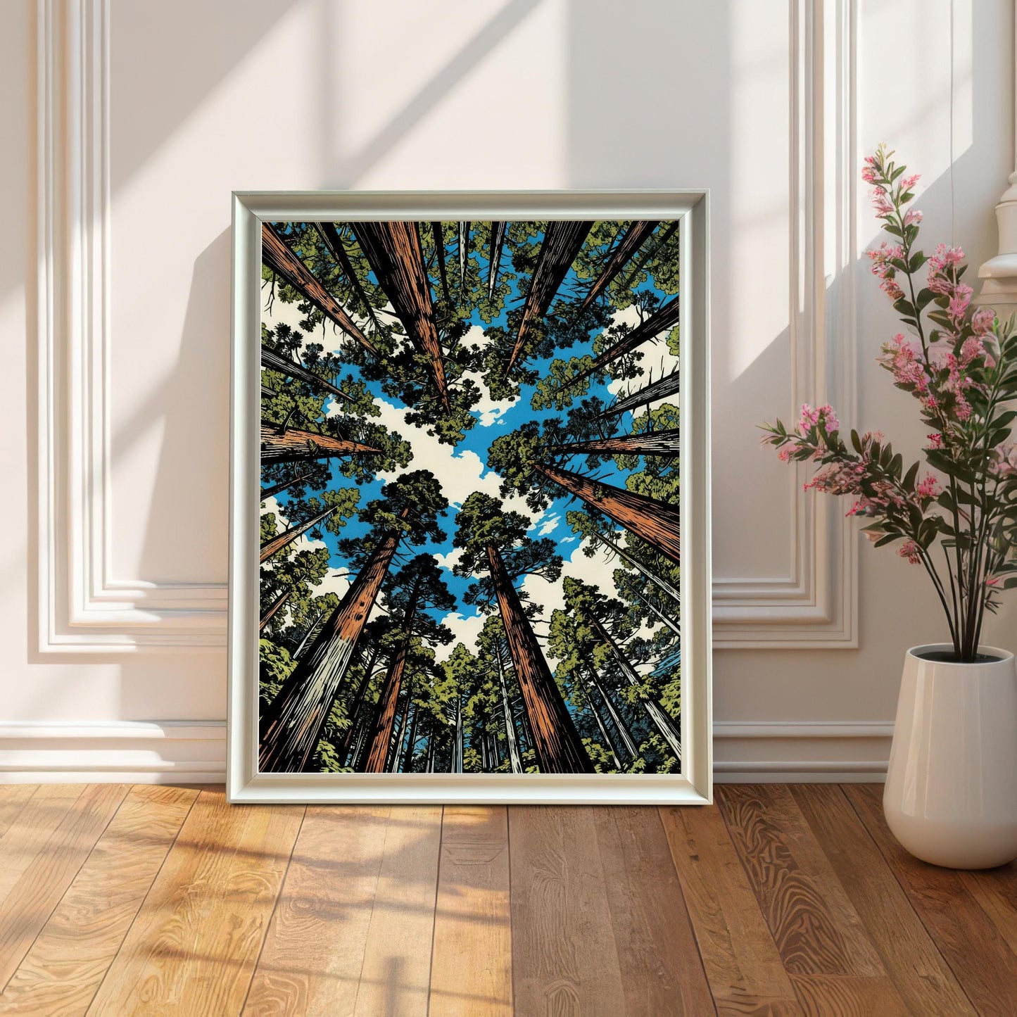 A white wood framed redwood forest print leaning against a decorative wall on a hardwood floor, styled next to a white vase with pink flowers.