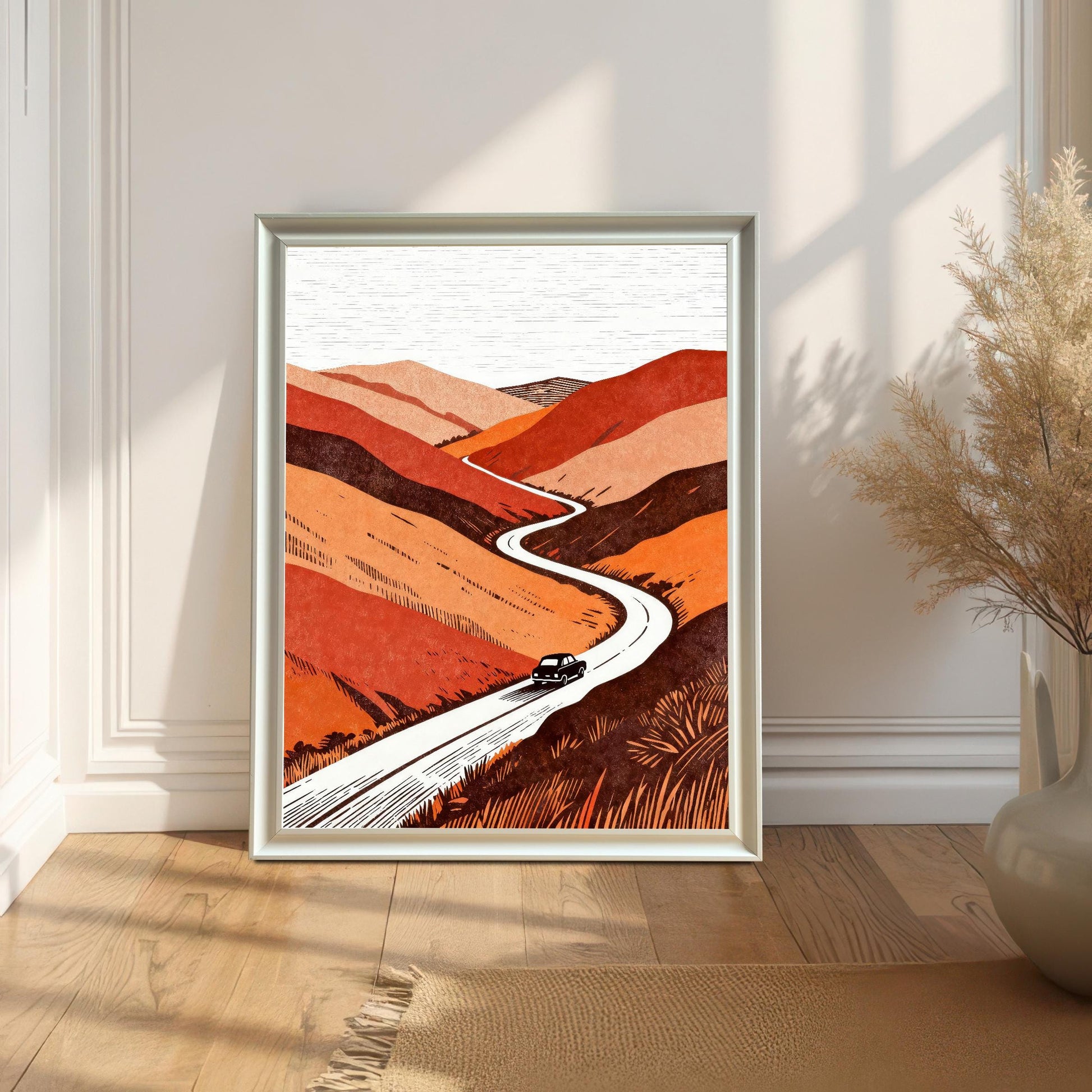 White-framed orange and earth-tone landscape print leaning against a white paneled wall on a light wood floor, captured in soft natural sunlight.