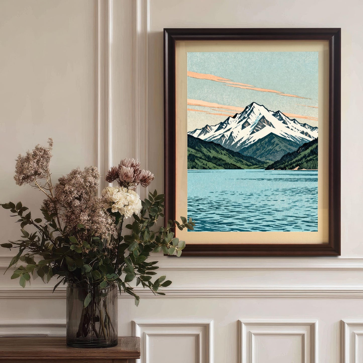 The mountain and lake art print in a dark coffee-colored wood frame with a mount, hanging on a classic white paneled wall above a vase of dried flowers.