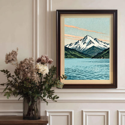 The mountain and lake art print in a dark coffee-colored wood frame with a mount, hanging on a classic white paneled wall above a vase of dried flowers.