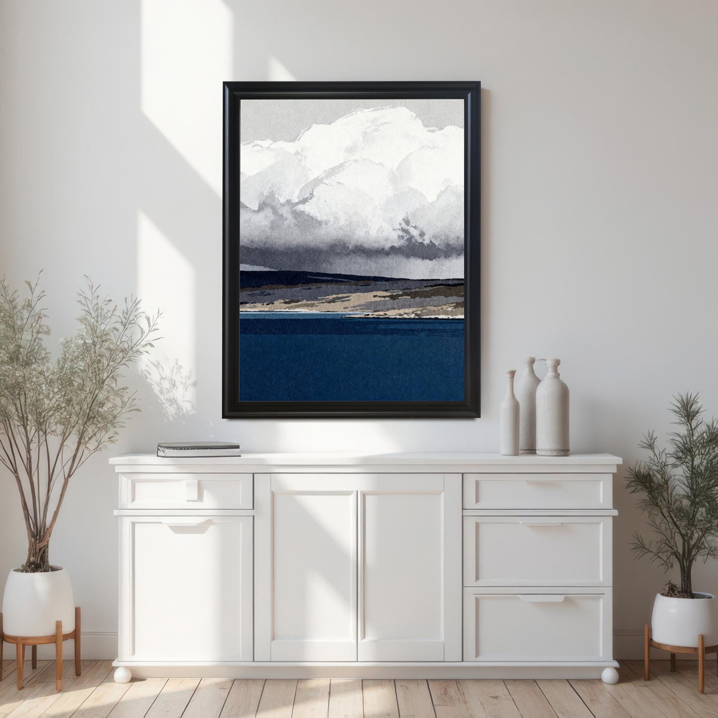 A black wood-framed abstract print of a navy blue sea and cloudy sky hanging on a white wall above a modern white sideboard, flanked by two potted plants in a bright, minimalist room.