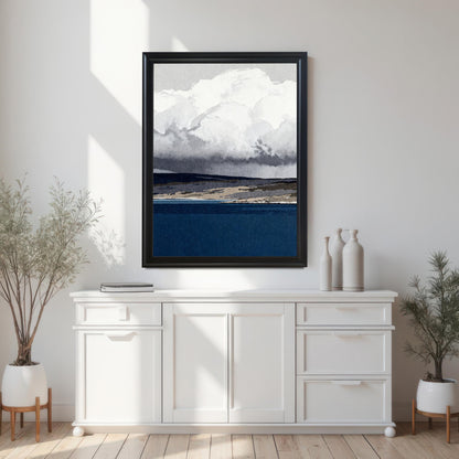 A black wood-framed abstract print of a navy blue sea and cloudy sky hanging on a white wall above a modern white sideboard, flanked by two potted plants in a bright, minimalist room.