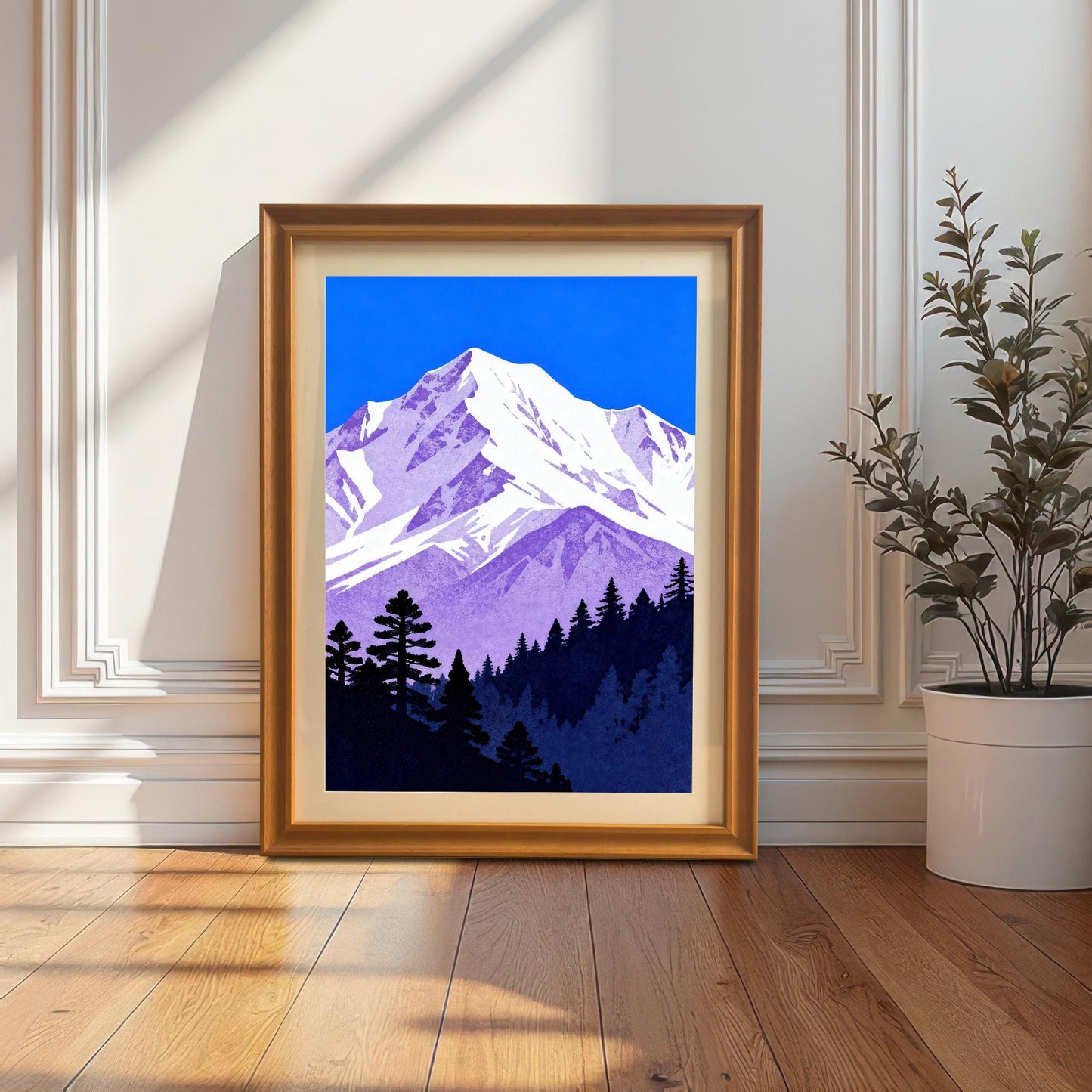 The purple and blue mountain art print in a gold-colored 'Orange' wood frame with a cream mount, leaning on a wooden floor against a white paneled wall.