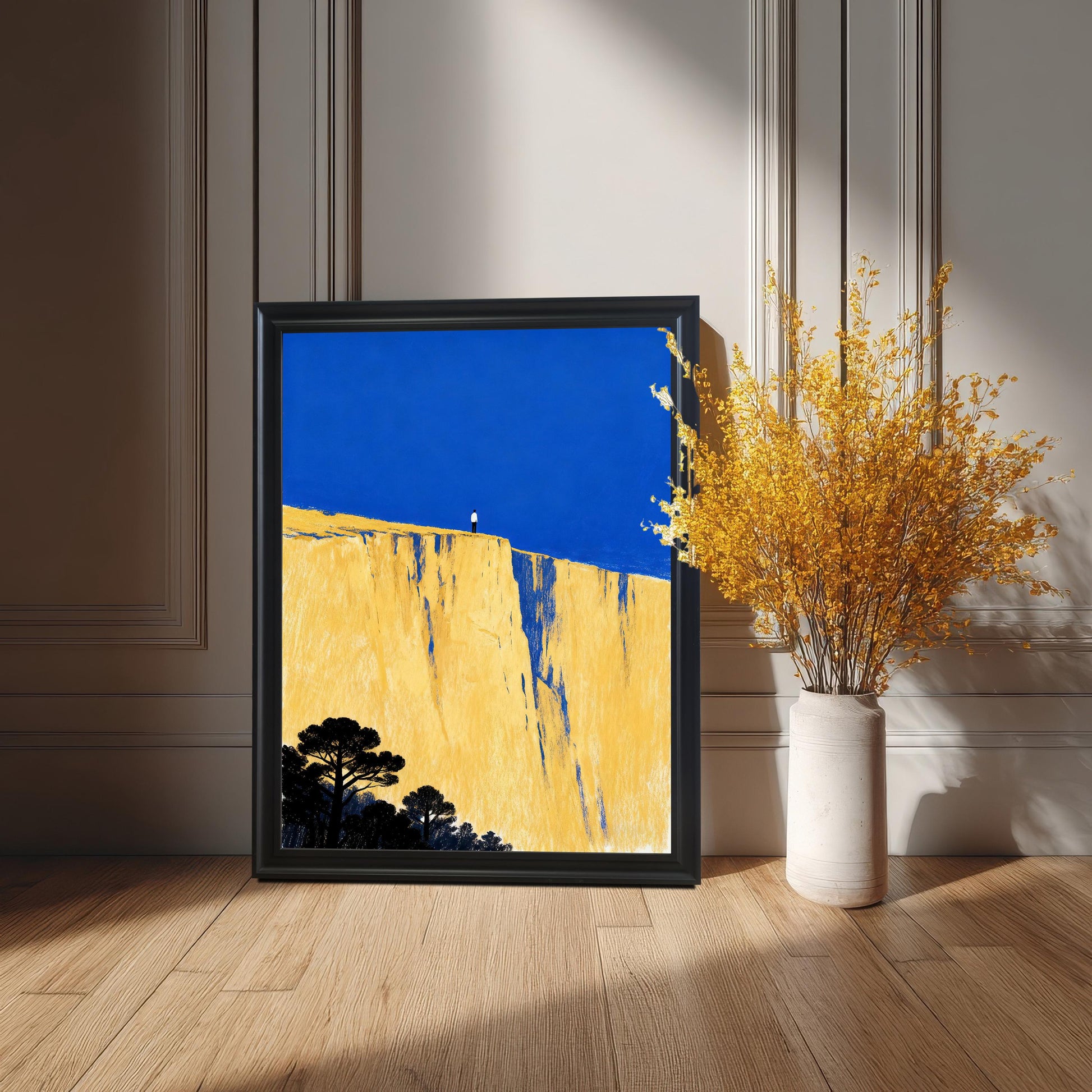 The art print in a black frame, resting on a light wood floor against a white paneled wall, beside a tall white vase filled with dried yellow flowers.