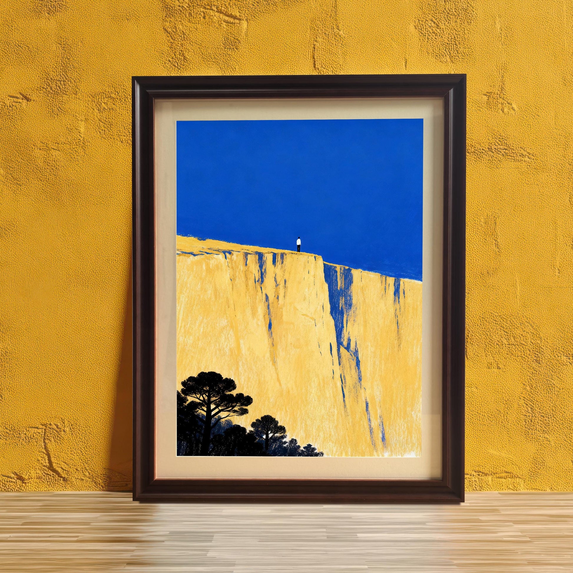 A framed art print featuring a figure on a yellow cliff edge beneath a bright blue sky, leaning against a textured mustard-yellow wall and a light wood floor. The frame is dark brown.