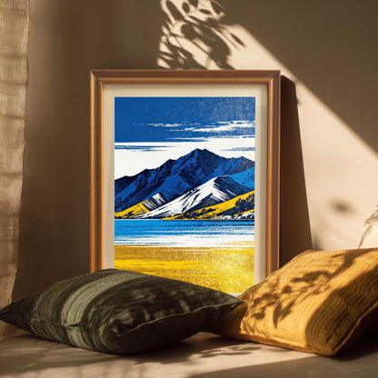 A framed art print in a light brown/orange wood frame, featuring the blue and yellow mountain landscape, resting on a beige surface with dark green and gold pillows. Natural light and leaf shadows create a moody atmosphere.