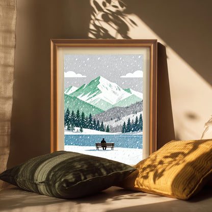 A framed art print in an orange wood frame, set on a cushioned surface with two pillows in sunlight. The artwork is a winter landscape with green mountains and a lone person sitting on a bench.
