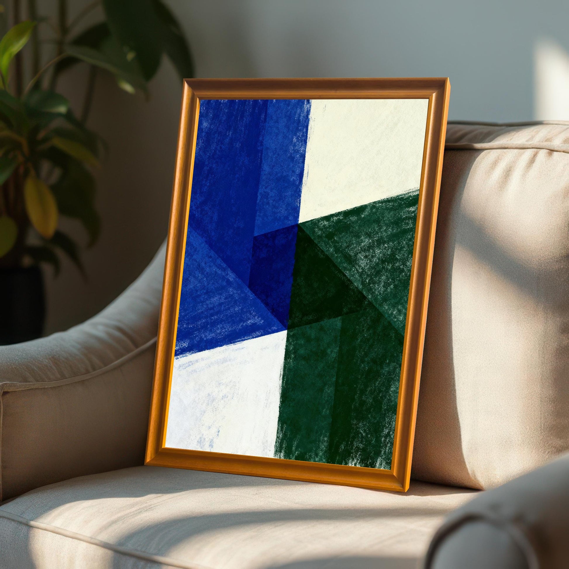 Orange-framed abstract art print leaning on a plush, light beige armchair or sofa in a well-lit room. The artwork is a modern geometric design in crayon texture with deep blue, dark green, and white/cream colors.