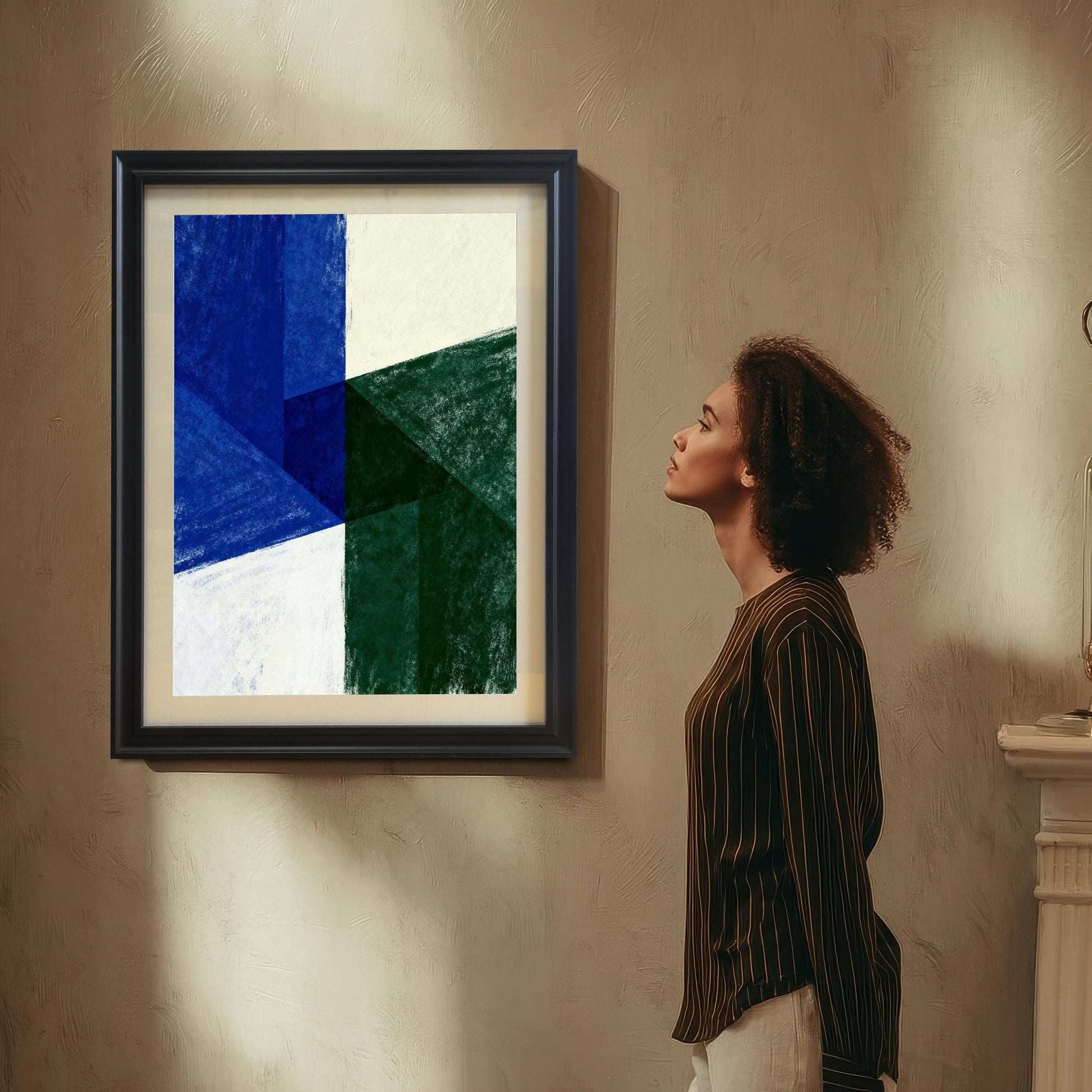 A woman with curly hair, wearing a striped shirt, standing in profile and looking up at the framed abstract art print hanging on a textured beige wall. The artwork is a collage of geometric shapes in deep blue, emerald green, and cream white.