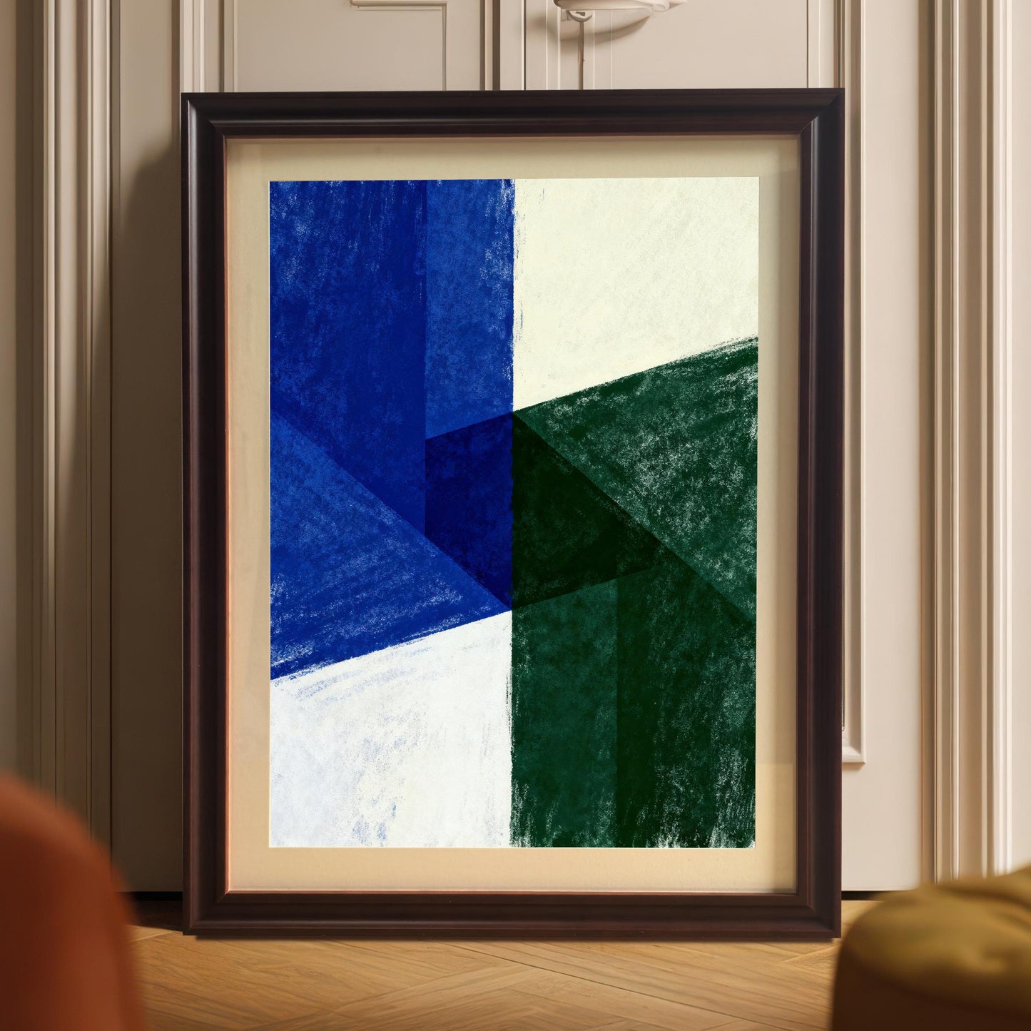 Dark Coffee-framed abstract art print with a mat, leaning on a wooden floor against white doors or a closet. The artwork is a bold geometric pattern in blue, green, and white/cream. Blurred orange and yellow furniture is visible in the foreground.