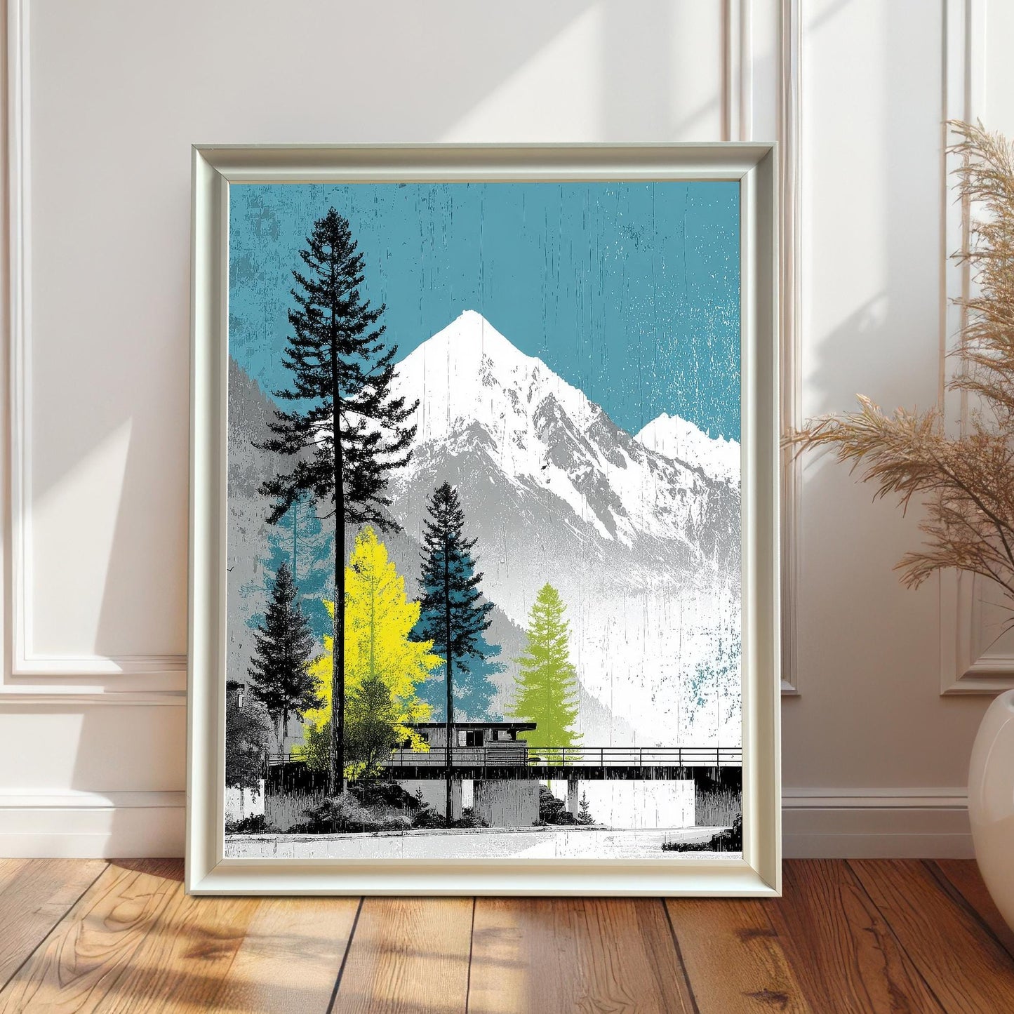 A white wood framed mountain landscape art print standing on a natural wood floor against a bright, modern white wall with decorative molding.