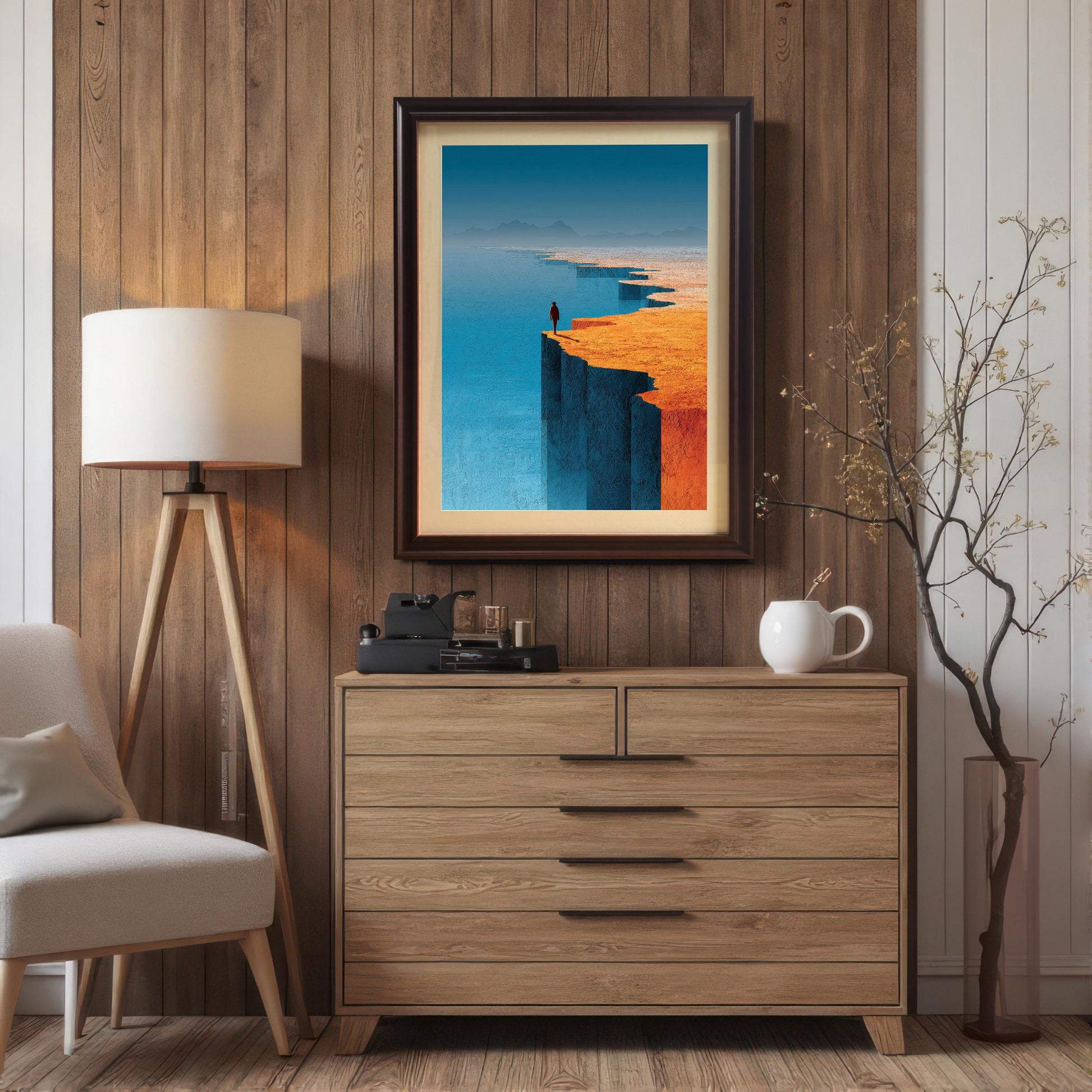 A landscape art print in a dark coffee wood frame with an off-white mat, hung on a rustic vertical-plank wood wall. The room setup includes a modern wooden dresser, a tripod floor lamp, and a decorative branch in a vase, creating a warm, contemporary interior.