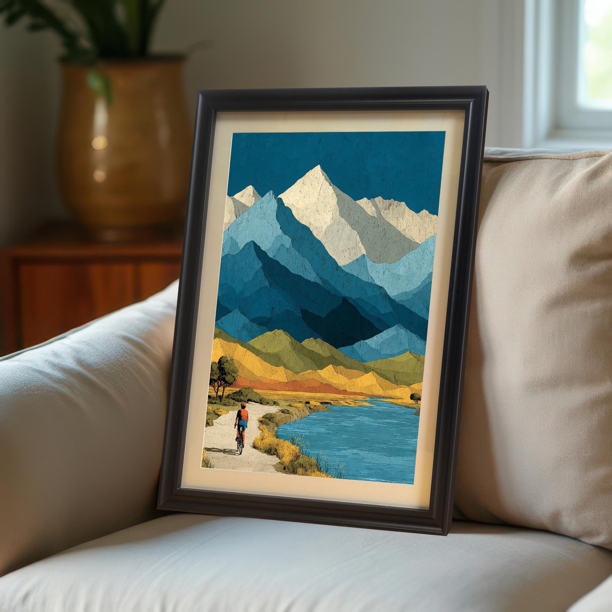A vertical framed art print of a stylized mountain landscape with a cyclist, leaning against a neutral-colored sofa in a bright, modern living room.
