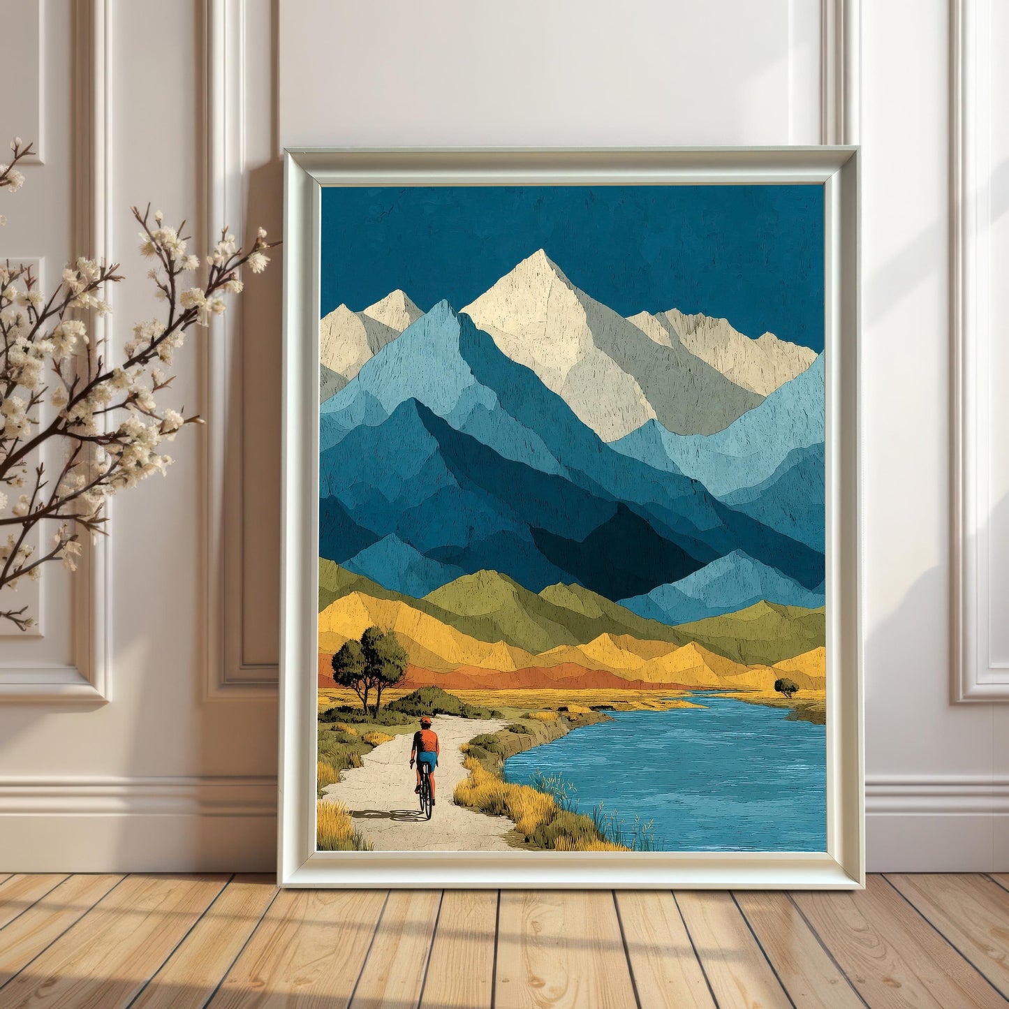 A large, white-framed mountain landscape art print leaning against a white paneled wall on a light wood floor, featuring a stylized scene of a cyclist near a blue lake.