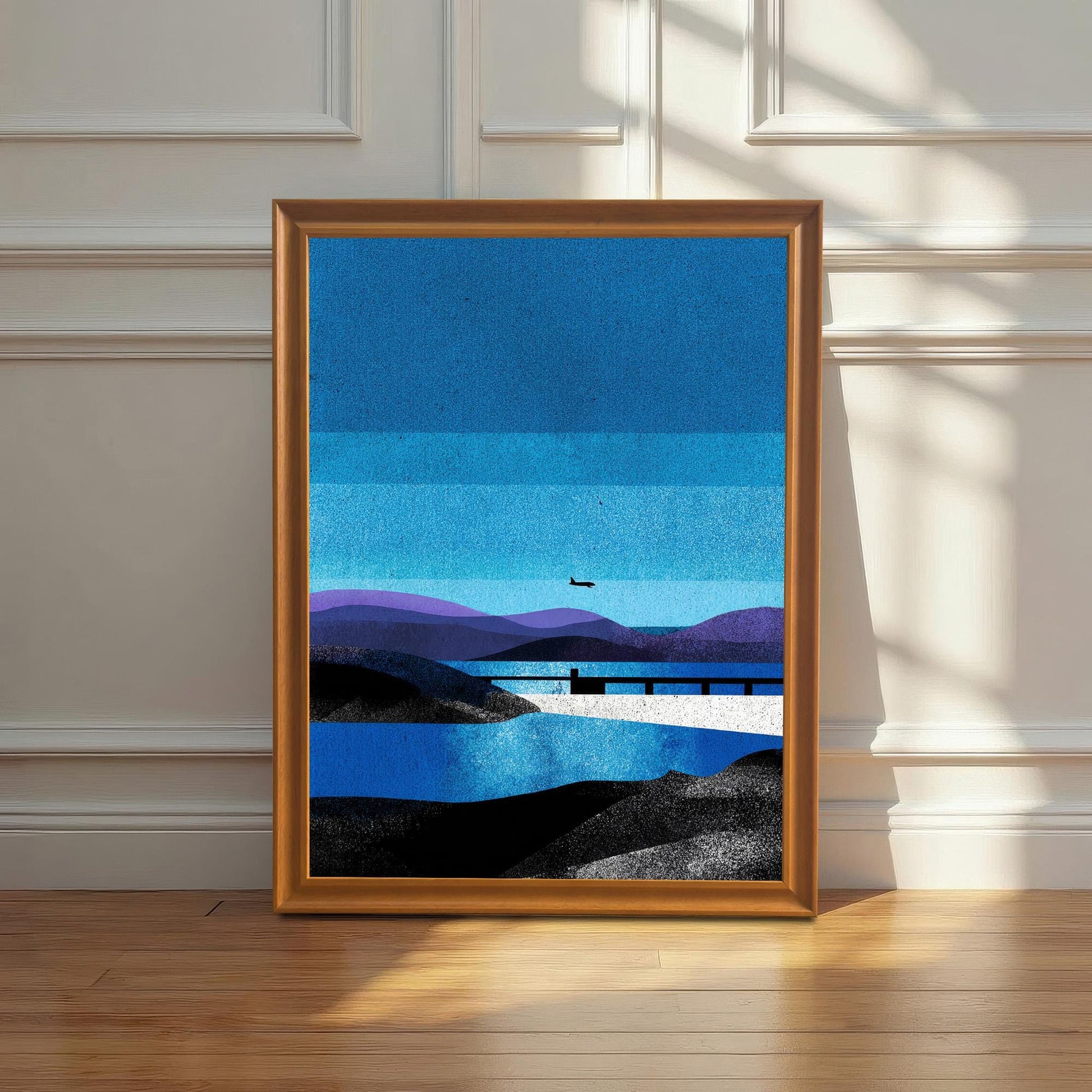A "Orange" wood-framed landscape art print leaning against a white wainscoted wall on a polished hardwood floor, illuminated by soft, natural morning sunlight.