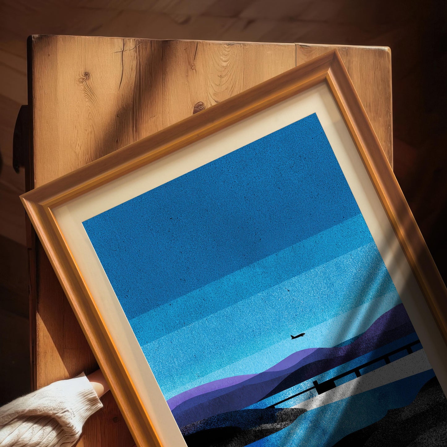 A high-angle detail shot of a hand holding the "Orange" wood-framed art print with an off-white mount, resting on a rustic wooden surface with natural sunlight and shadows.