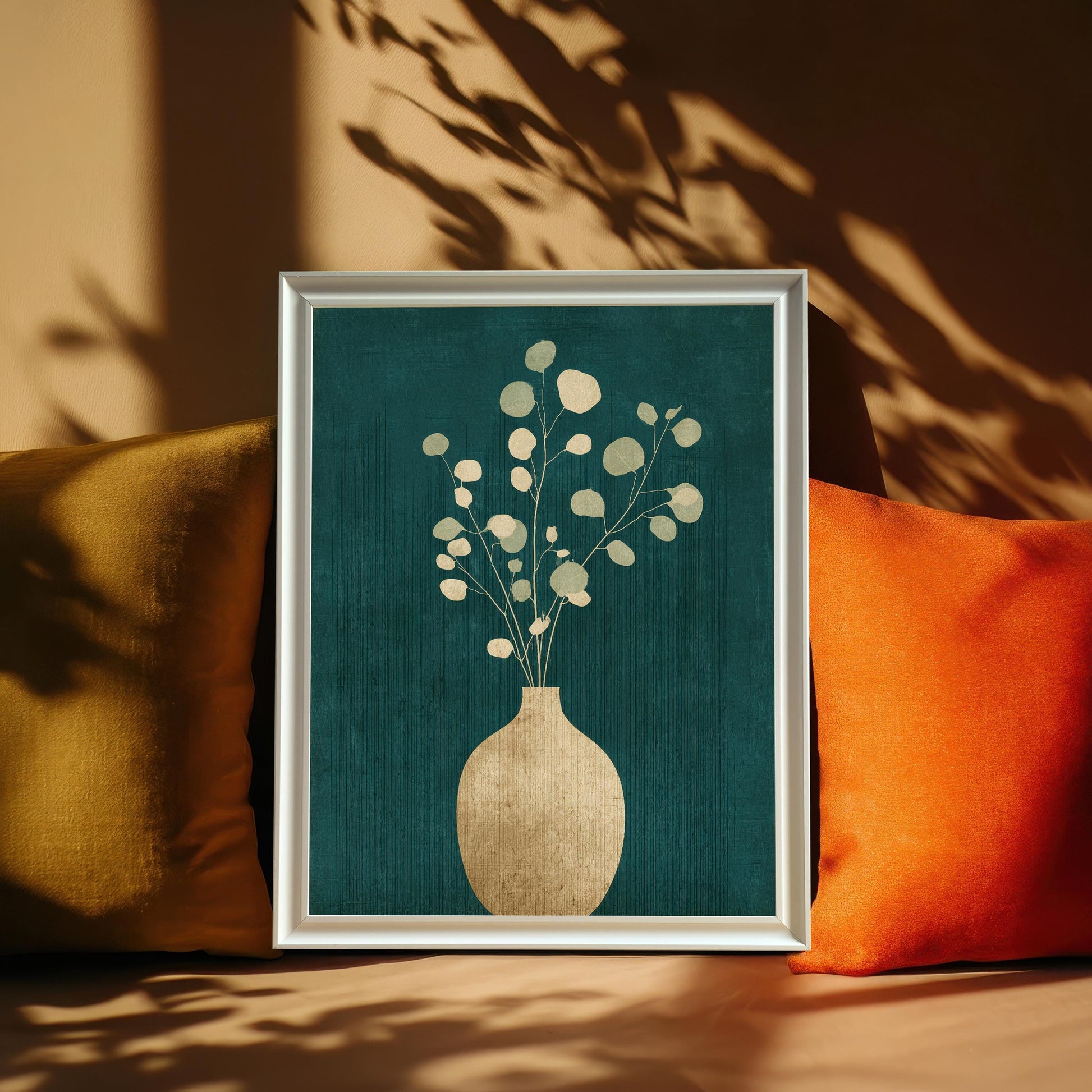 A framed art print featuring a gold vase and eucalyptus leaves on a dark teal background, leaning against a warm beige wall between yellow and orange pillows with artistic leaf shadows.