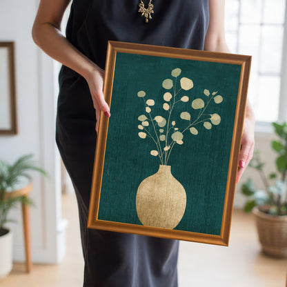 A person in a black dress holding a large, orange-toned wood-framed art print of a gold eucalyptus plant to demonstrate the scale and style of the piece.