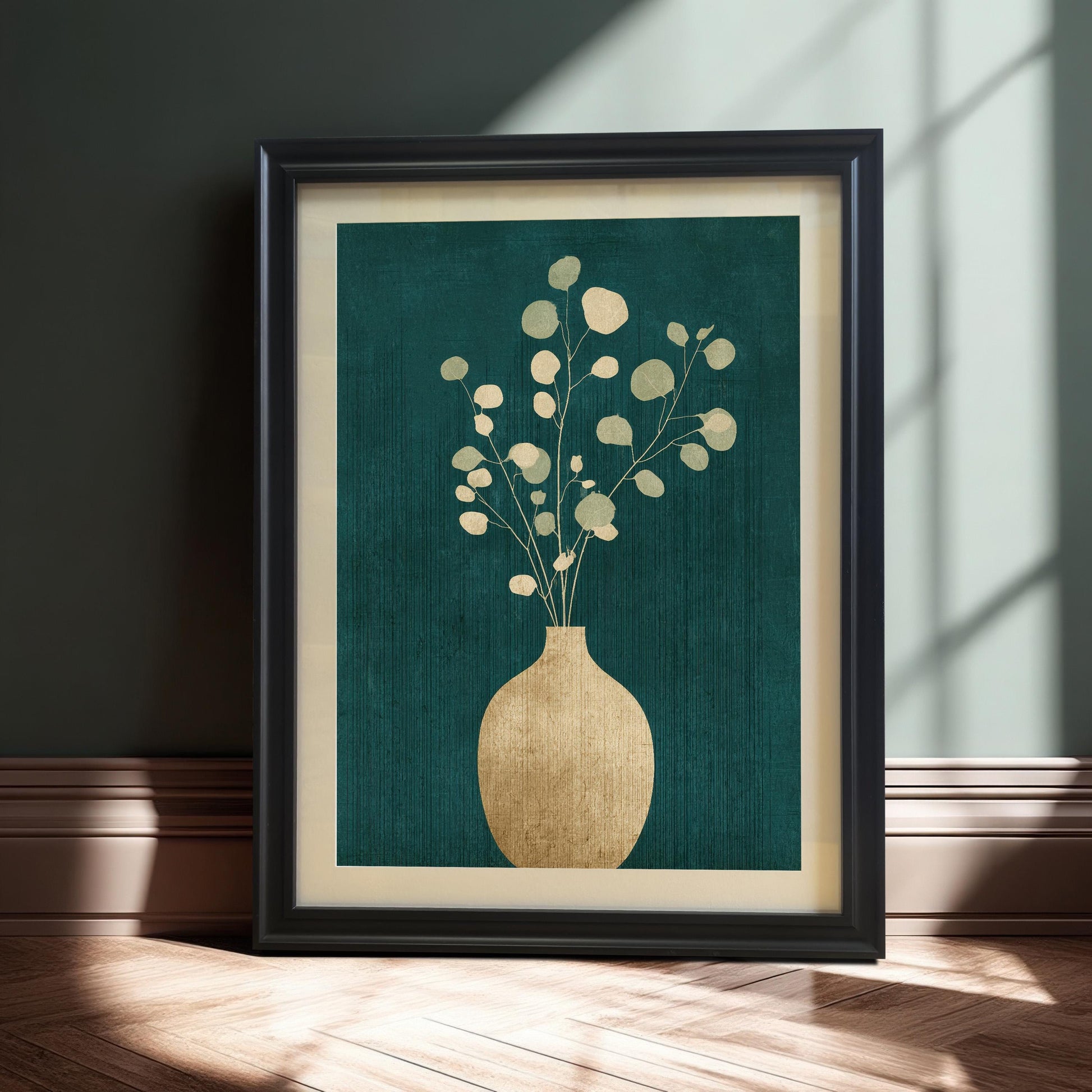 A black-framed botanical art print featuring a golden vase and eucalyptus branches on a textured teal background, leaning against a dark wall on a light wood floor with soft natural light shadows.