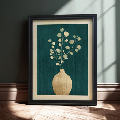 A black-framed botanical art print featuring a golden vase and eucalyptus branches on a textured teal background, leaning against a dark wall on a light wood floor with soft natural light shadows.