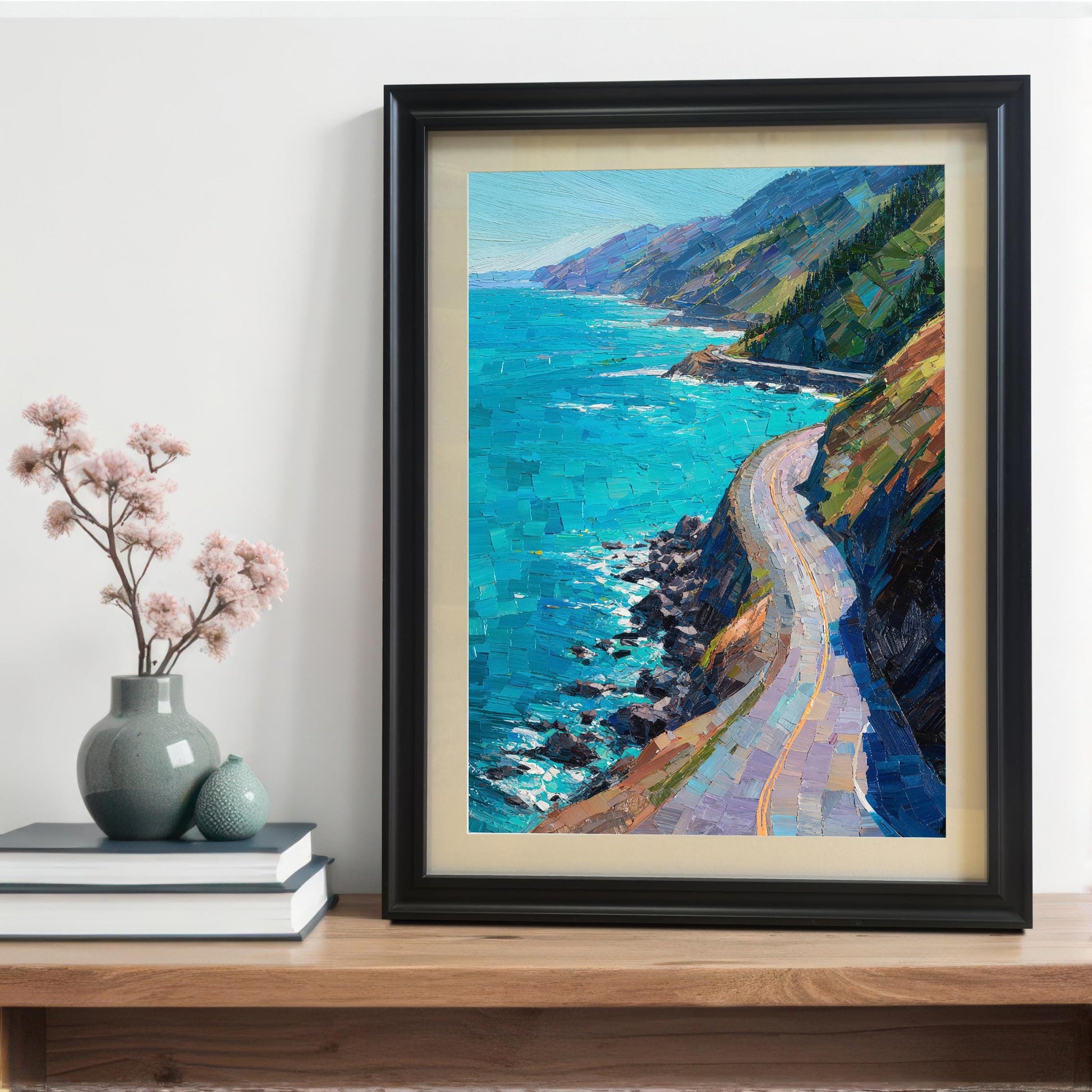 A black wood framed coastal art print with a cream mat board, leaning on a wooden tabletop next to a stack of books and a small gray vase with pink dried flowers.