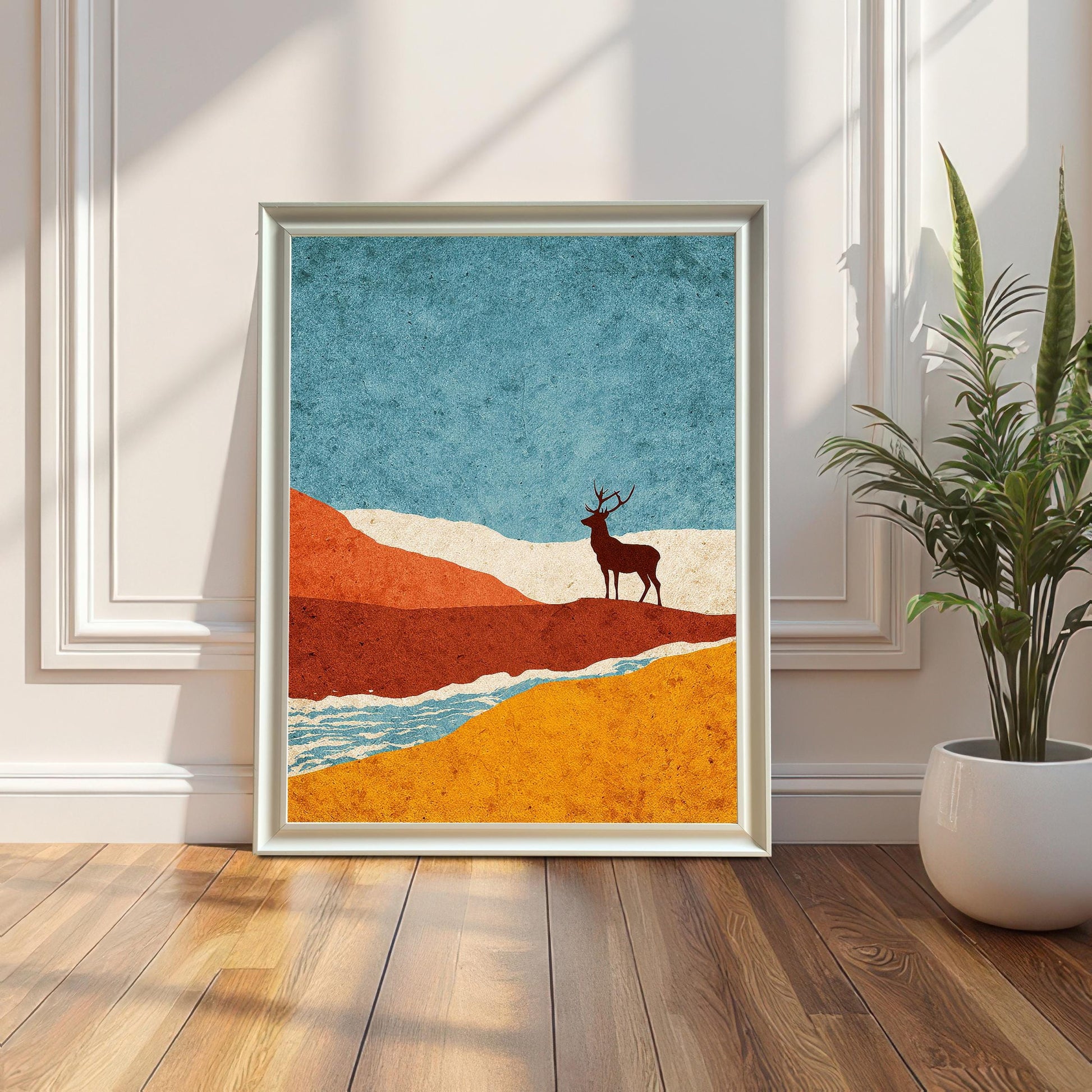 A large white-framed minimalist art print featuring a stag silhouette in a landscape of teal, orange, and gold, leaning against a white paneled wall next to a potted plant.