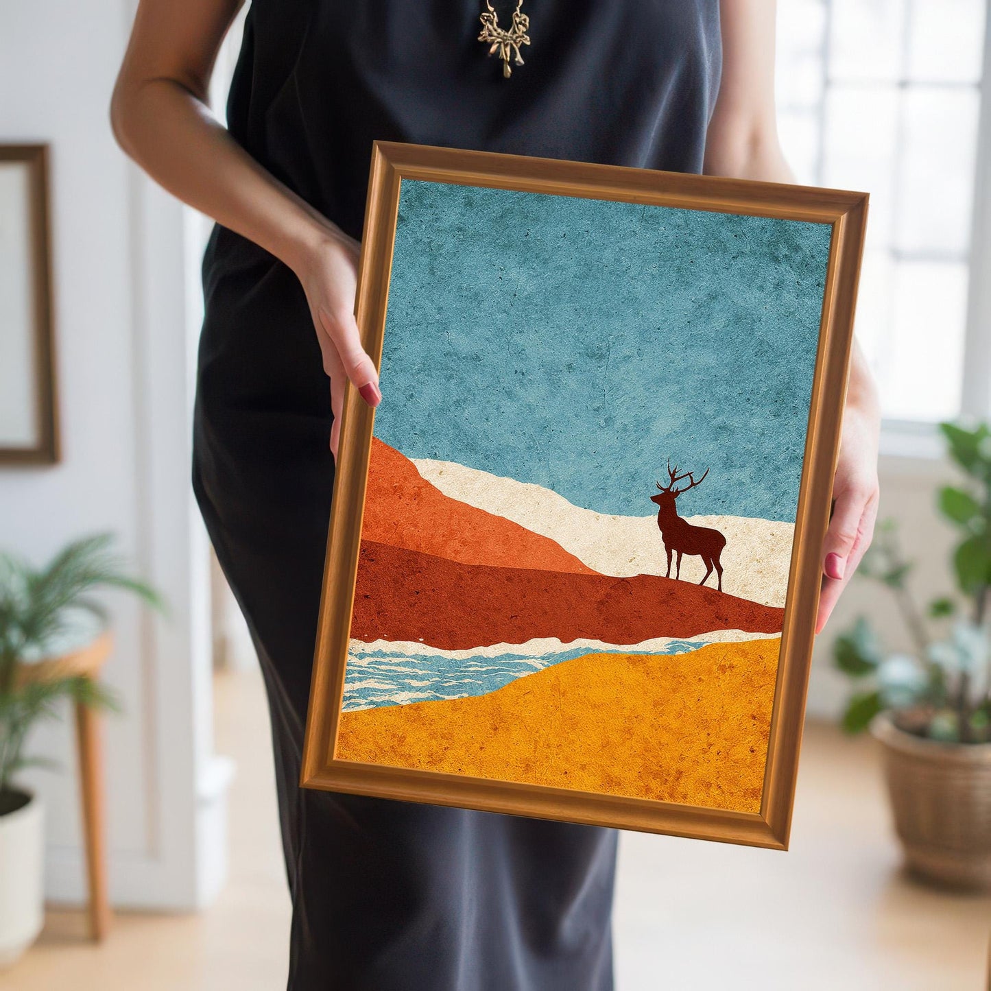 A person holding the orange-framed stag art print to provide a real-world scale and showing the vibrant colors of the mid-century modern style landscape.