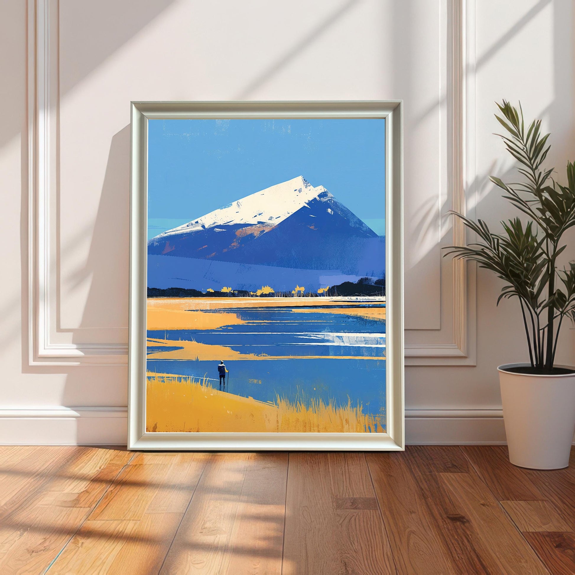 A framed art print of a snowy mountain peak overlooking a blue lake and golden fields, leaning against a white paneled wall on a light wood floor. To the right, a green potted plant sits in a white ceramic pot, with soft sunlight casting shadows across the scene.