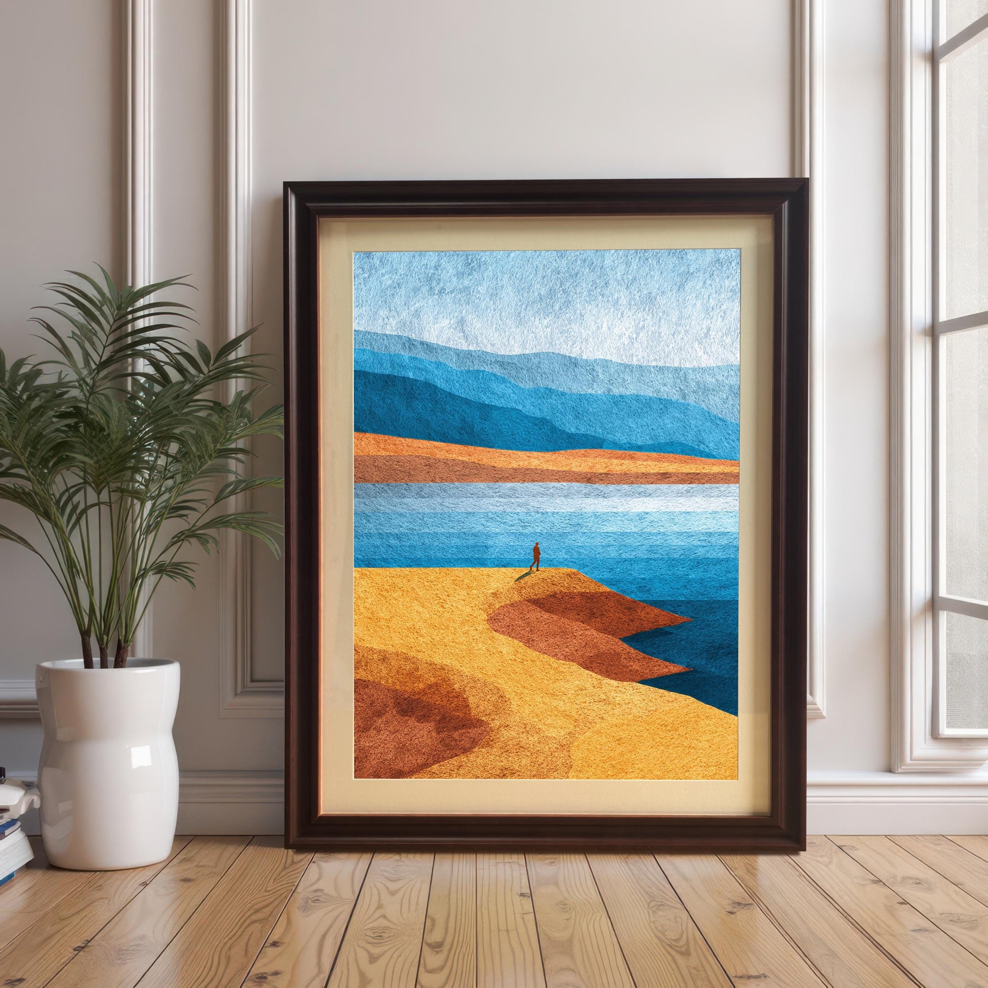 Minimalist landscape art print in a dark coffee wood frame with an off-white mat board, leaning against a white paneled wall next to a potted plant on hardwood floors.