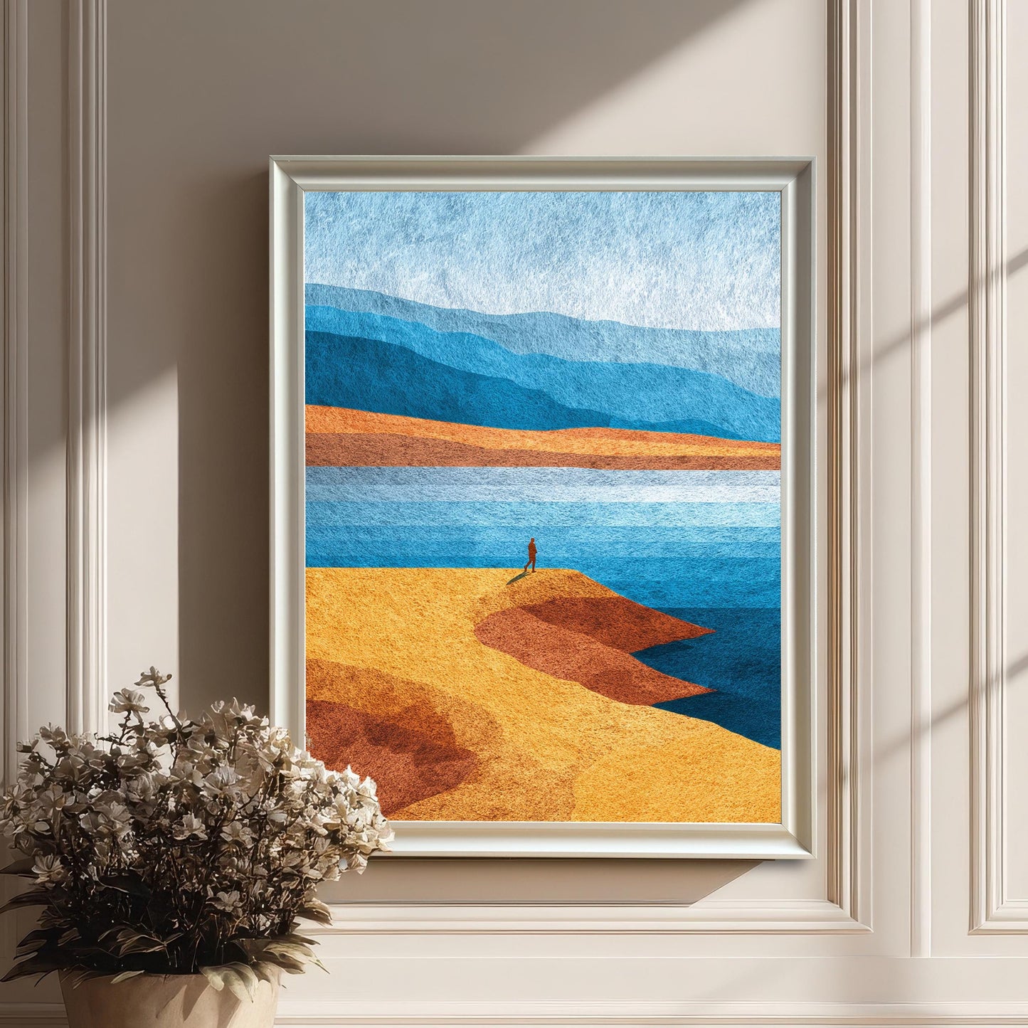 White wood framed abstract coastal art print mounted on a classic paneled wall, featuring a small white floral arrangement in a pot on the floor below.