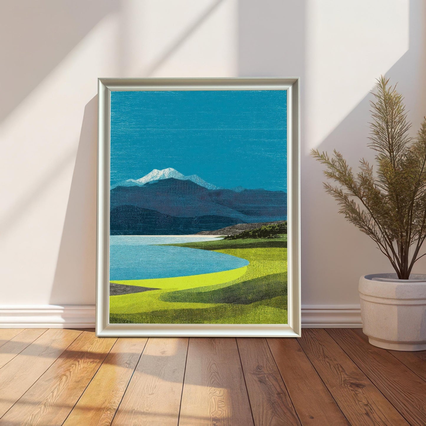 A vertical white-framed landscape art print of a blue lake and snow-capped mountain, leaning against a white wall on a wooden floor next to a potted plant.