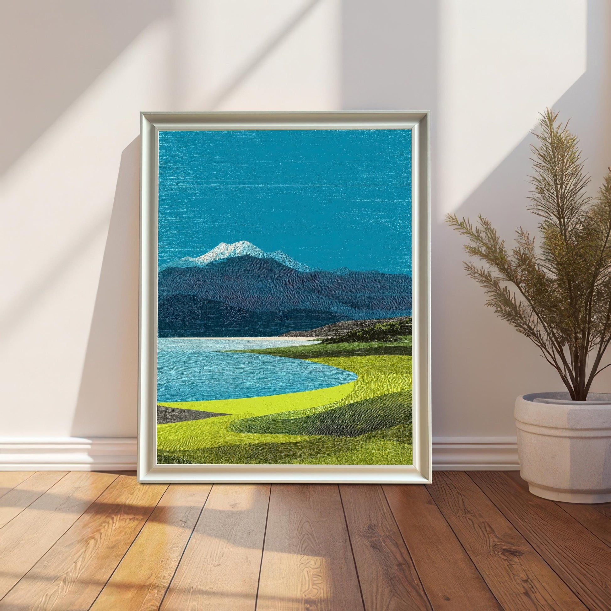 A vertical white-framed landscape art print of a blue lake and snow-capped mountain, leaning against a white wall on a wooden floor next to a potted plant.