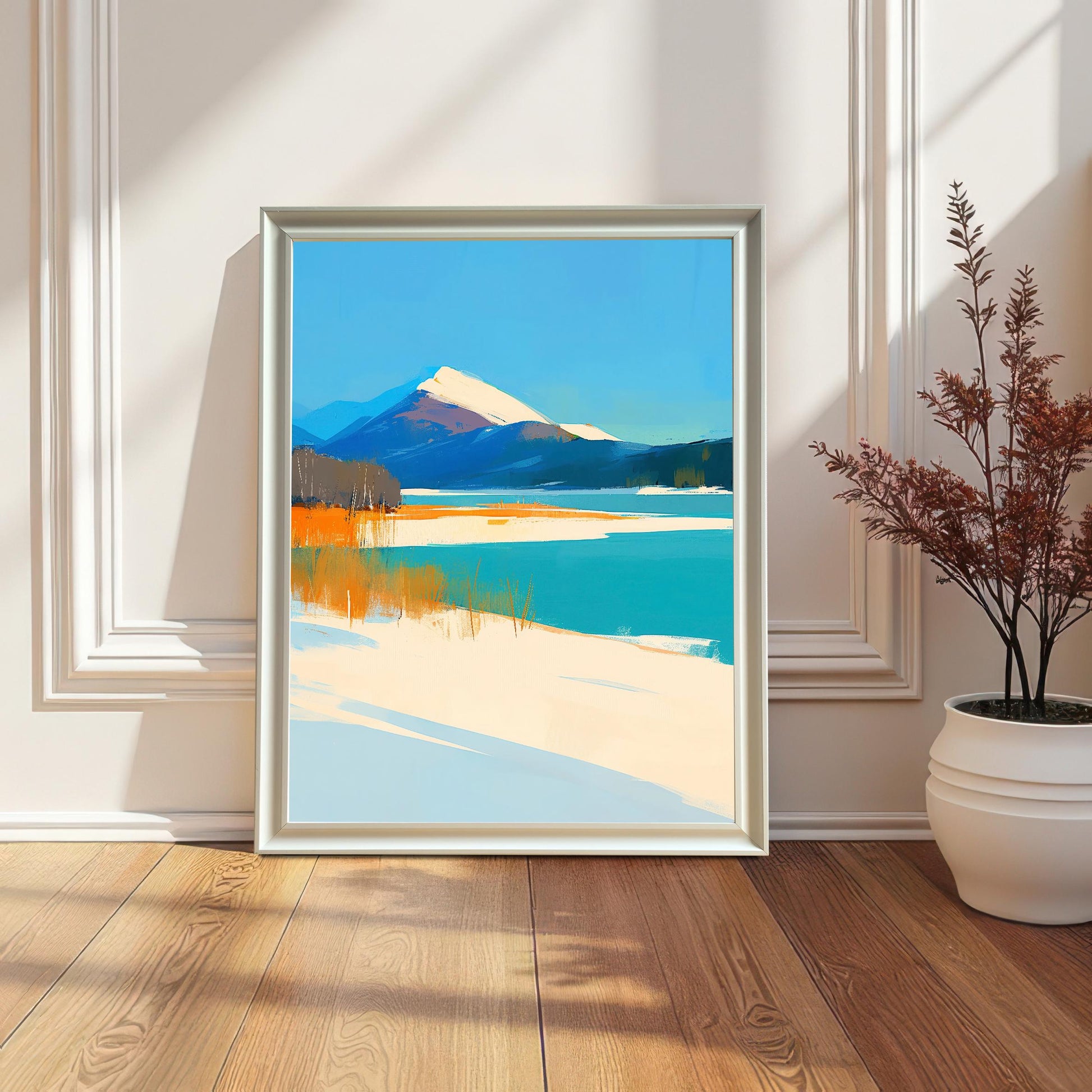 A vertical landscape art print with a white frame leaning against a cream-colored wall with decorative molding. The artwork features a snowy mountain and a turquoise lake. A white vase with dried branches sits to the right on a wooden floor.