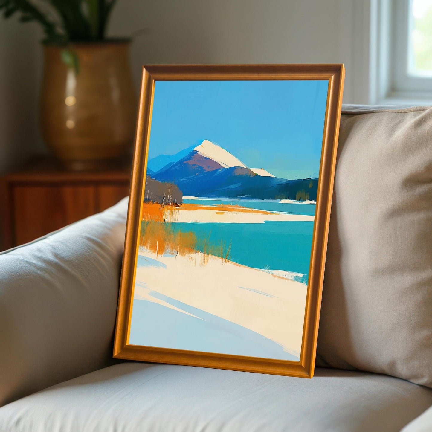 A lifestyle shot of the art print in an orange-toned wood frame resting on a light grey fabric sofa, showcasing the vibrant colors of the mountain landscape.