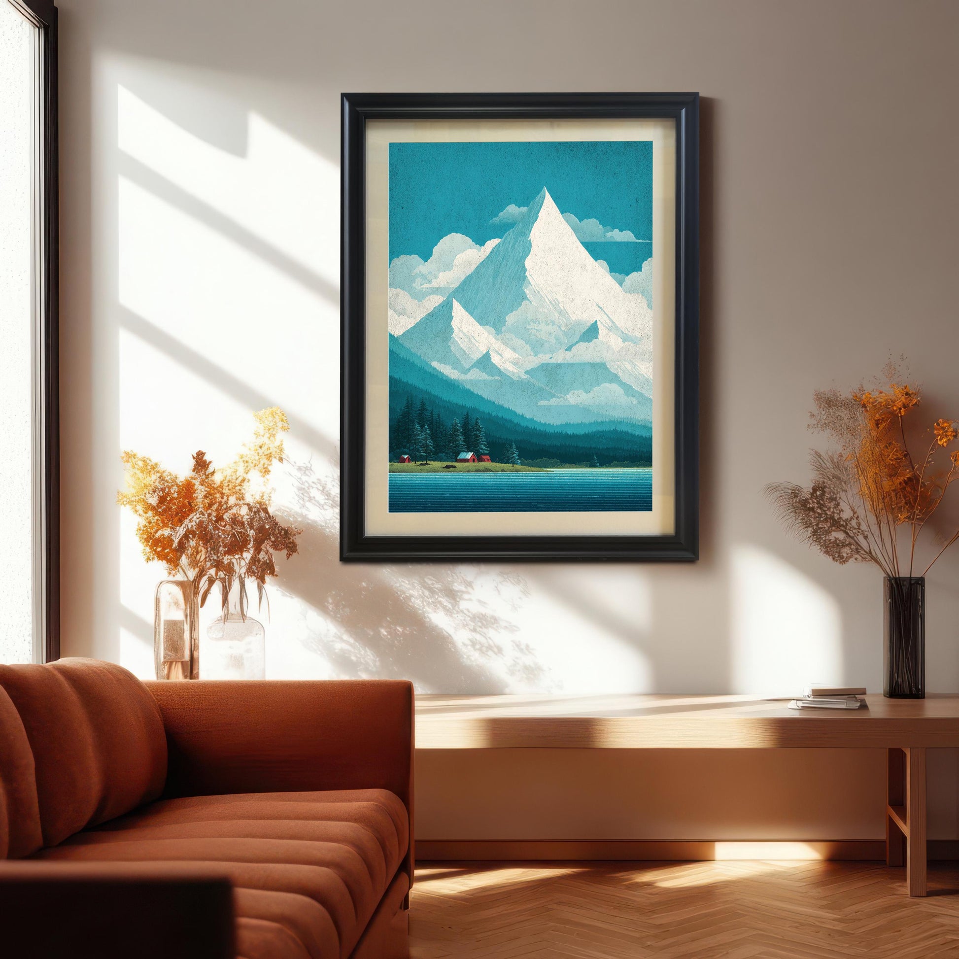 A black wood-framed mountain art print with a cream mat mount hanging in a bright, sunlit living room above a minimalist wooden bench and an orange velvet sofa.