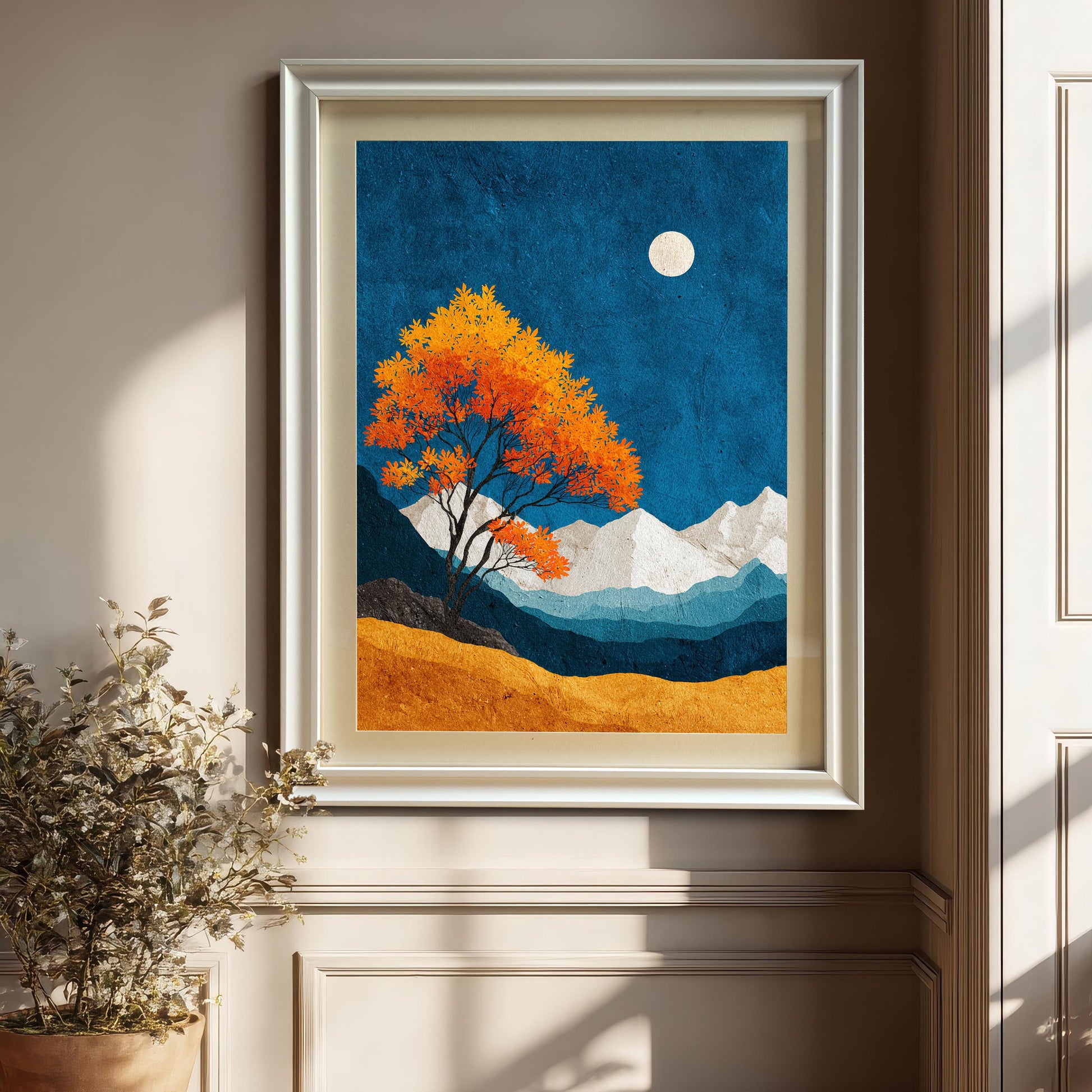 A modern white-framed art print of an orange autumn tree against a blue night sky, displayed on a neutral-colored wall above decorative wainscoting and a potted plant.